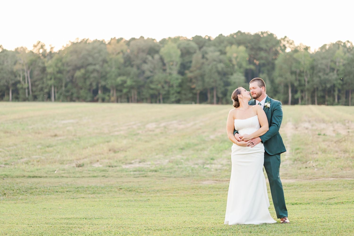 A couple shares a romantic dance in a sunlit meadow wearing formal wedding attire.