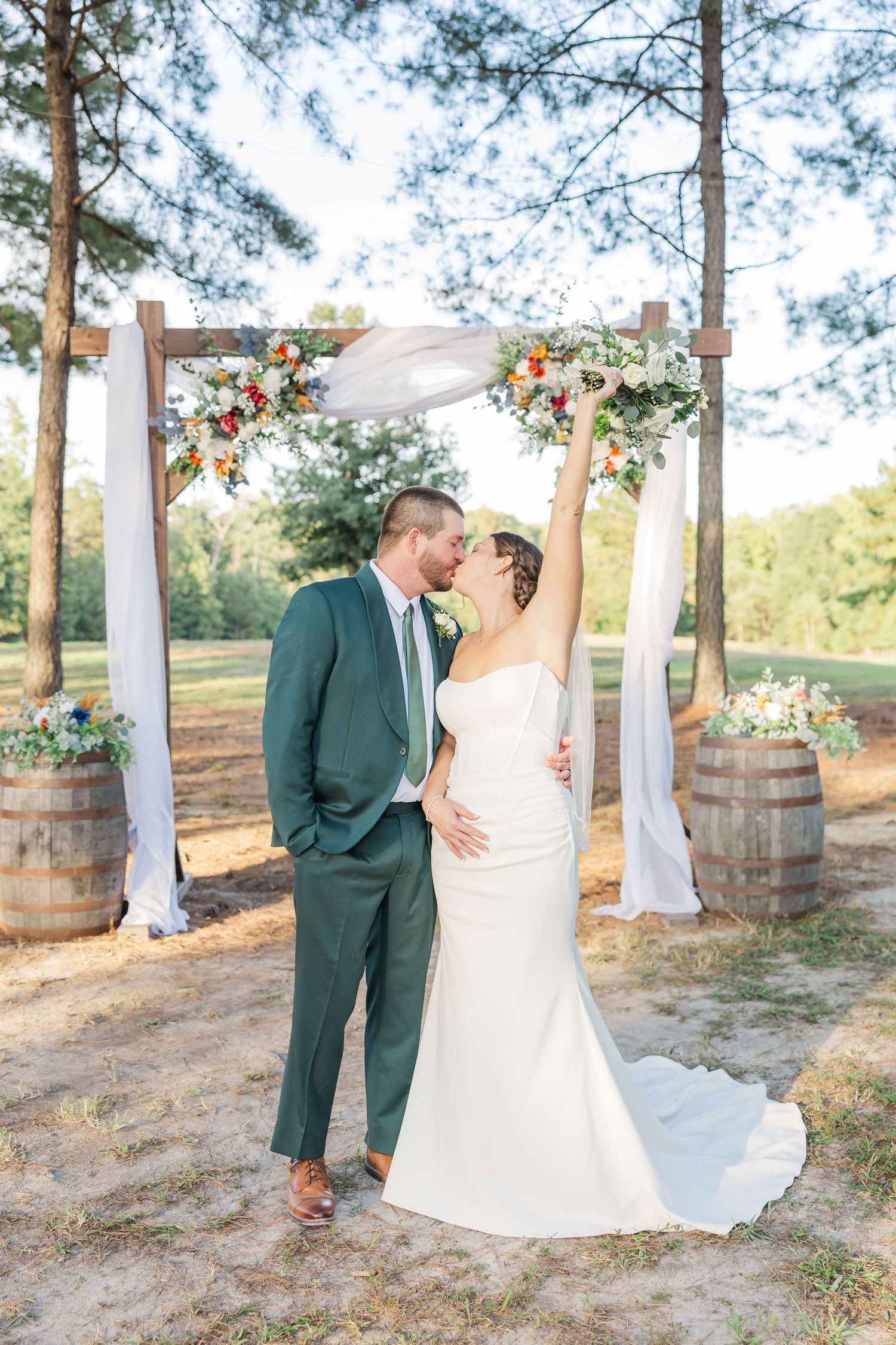 A wedding couple poses under a rustic wooden arch decorated with flowers and white fabric in a pine forest setting.