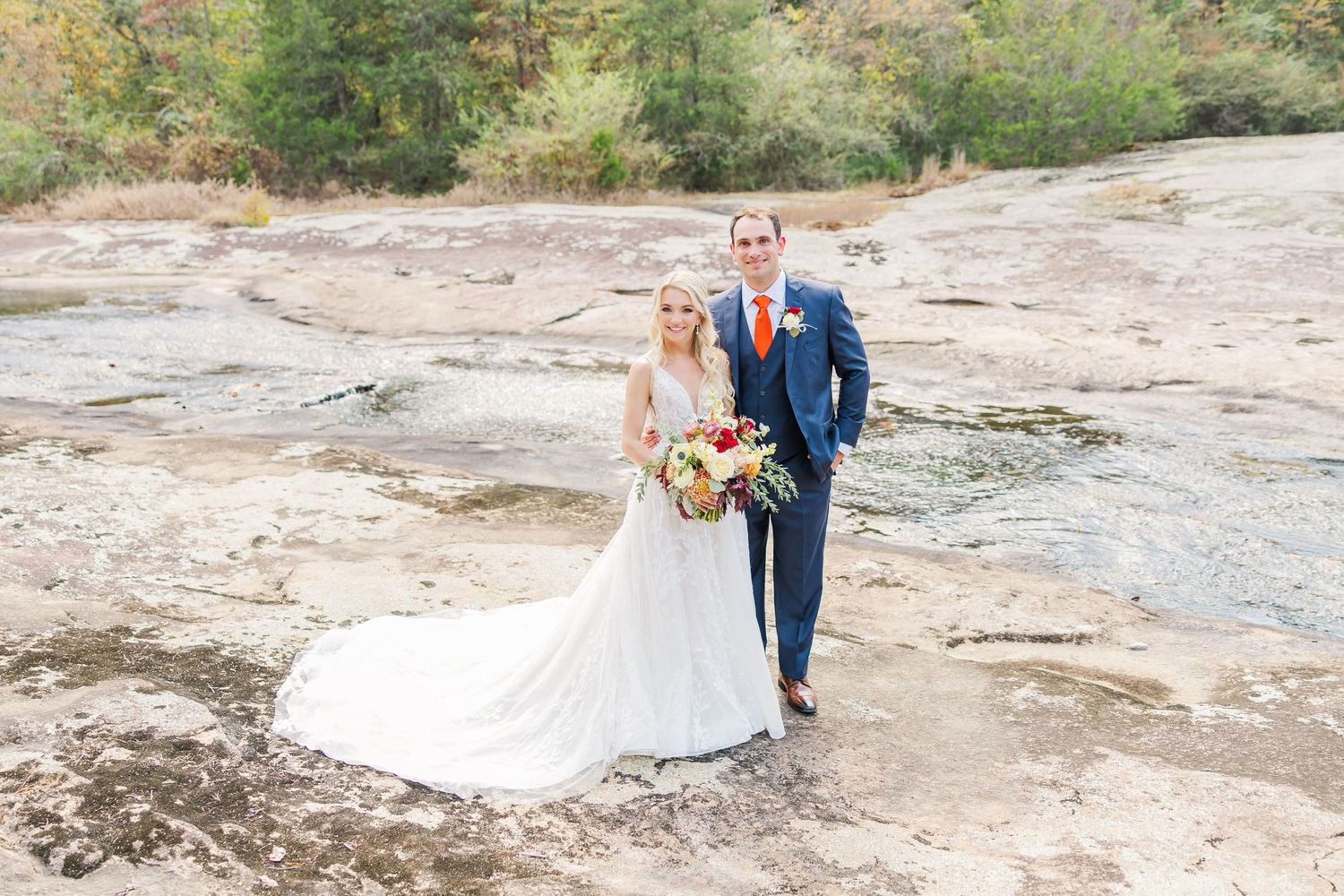 A couple poses for their wedding photos on rocky terrain wearing formal attire with a flowing white dress.