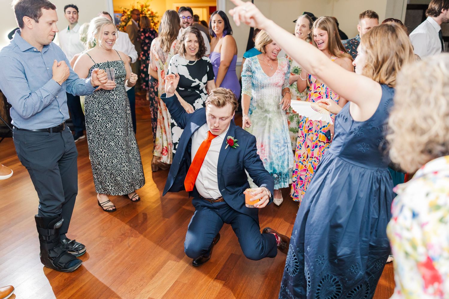 Wedding guests dance and celebrate at a reception on a hardwood floor as a guest in a red tie kneels in the center.