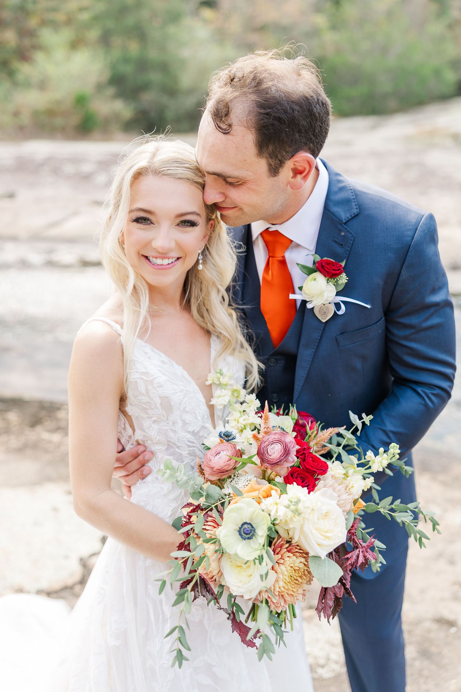 A romantic wedding photoshoot featuring a navy suit with orange tie and a beautiful floral bouquet with red and white flowers.