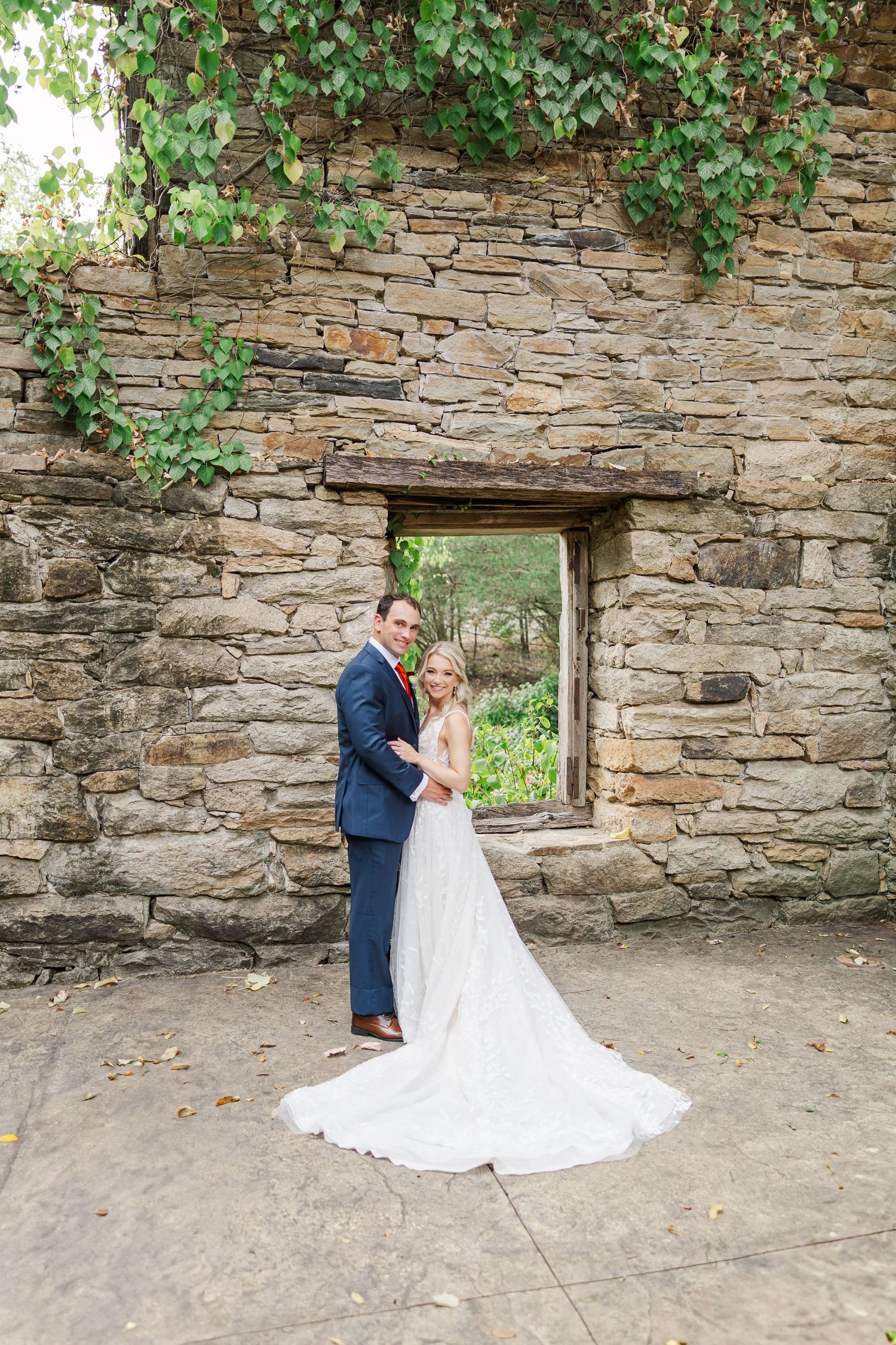 A romantic wedding portrait showing a couple embracing by an old stone wall with trailing ivy and a window opening.
