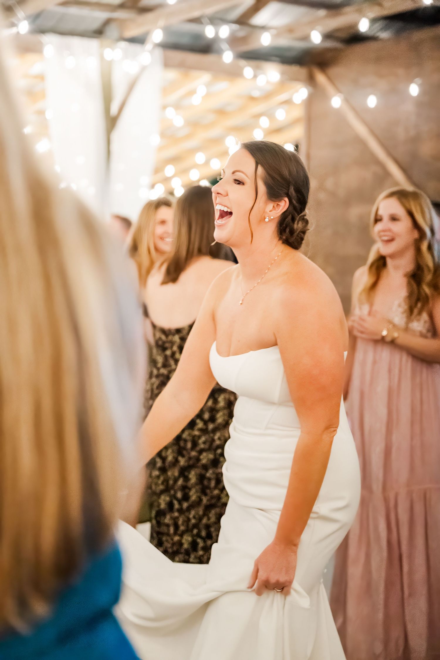Joyful bride in white strapless gown laughing at wedding reception with string lights and guests.