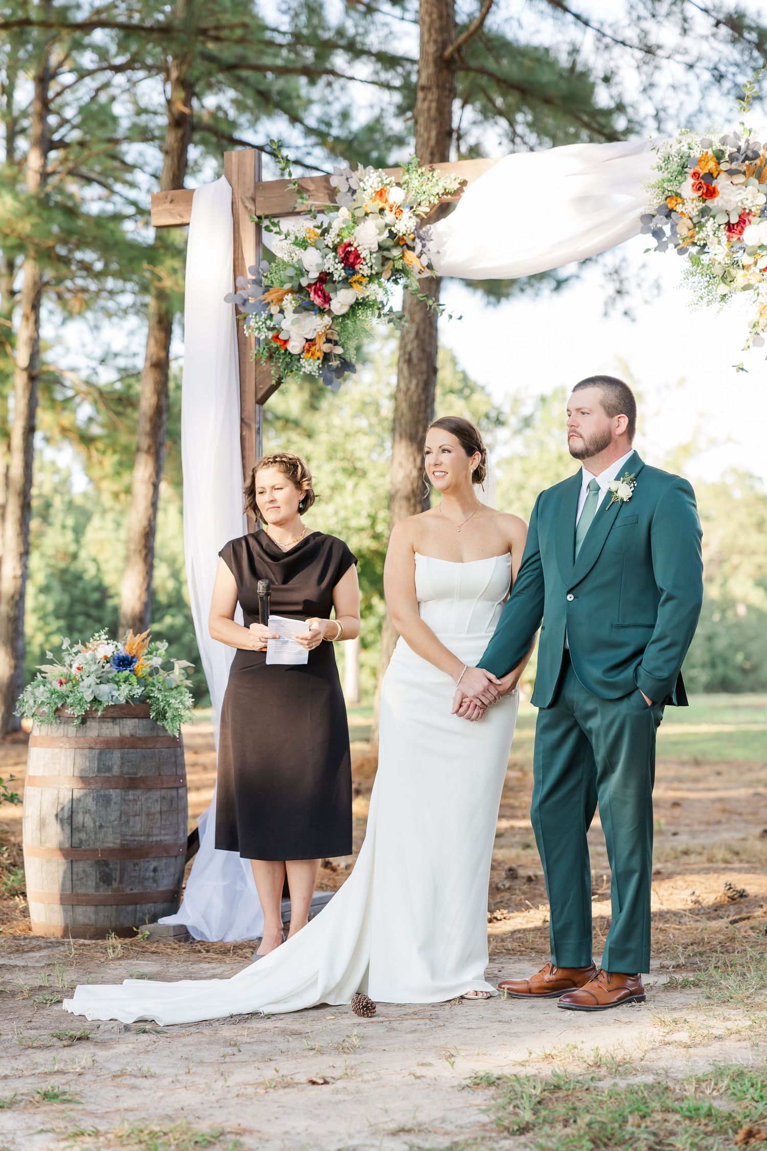 Bride and groom hold hands during outdoor wedding ceremony beneath rustic wooden arch with colorful floral arrangements.