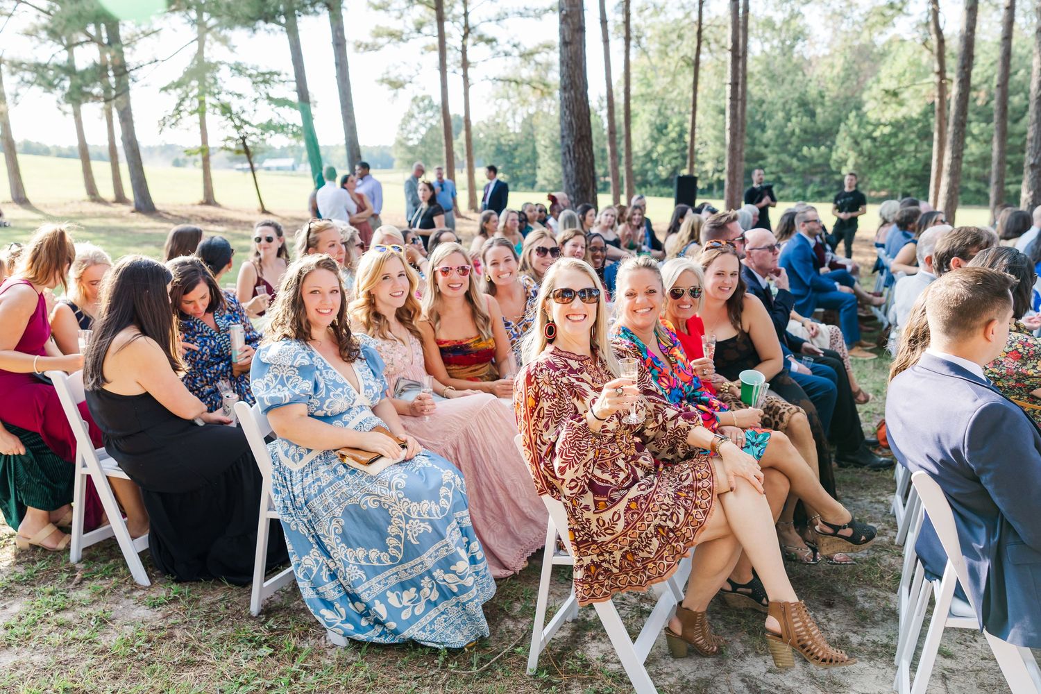Guests smiling and seated in white chairs at an outdoor wedding ceremony surrounded by tall pine trees.