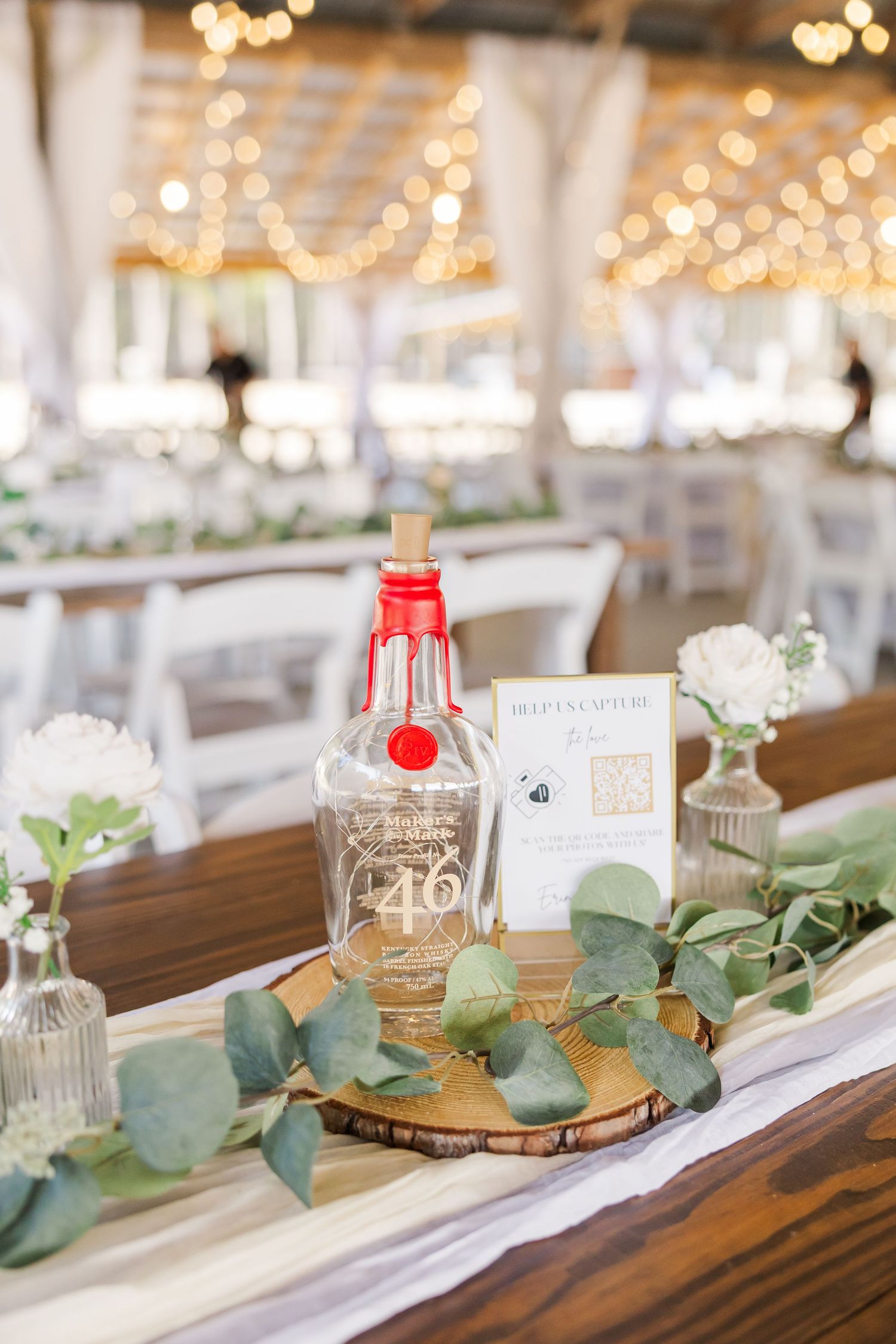 Wedding table centerpiece featuring eucalyptus greenery, white flowers, a decorative bottle on wood slice, and a photo capture sign.