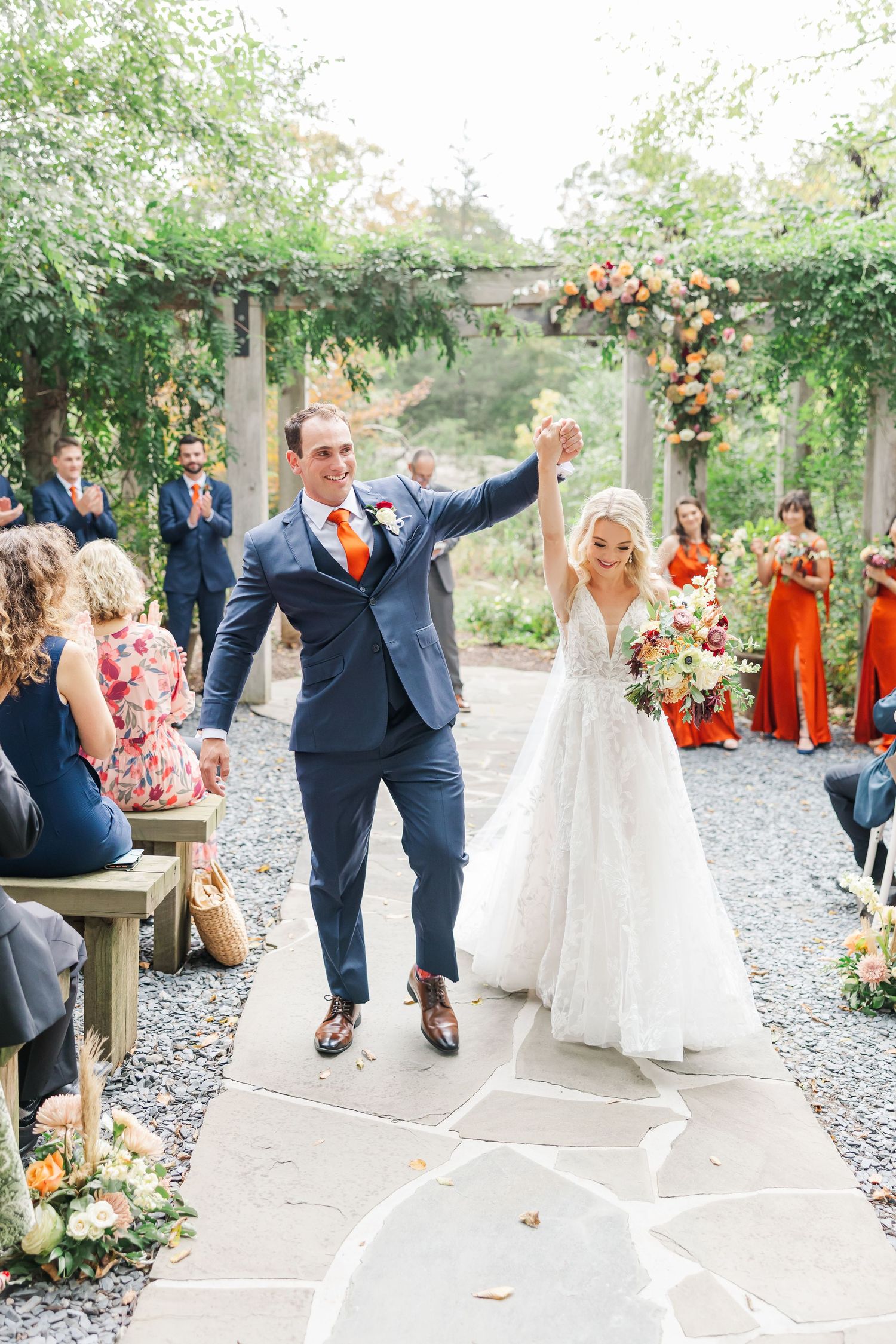 Joyful wedding celebration under garden pergola with gravel pathway and orange floral decorations.