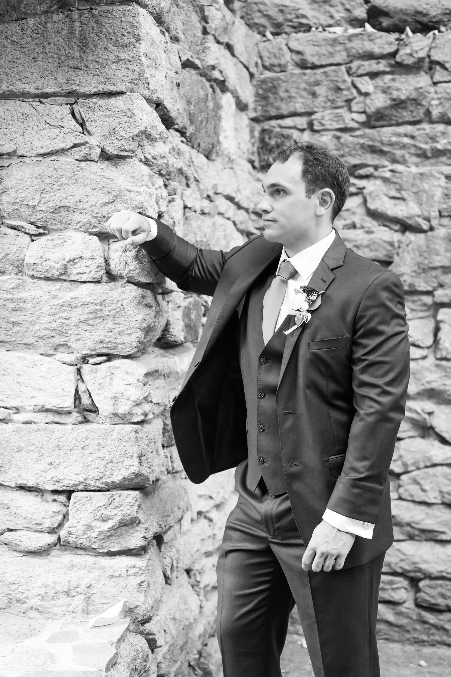 Two people in formal black suits pose playfully against a rustic stone wall backdrop.