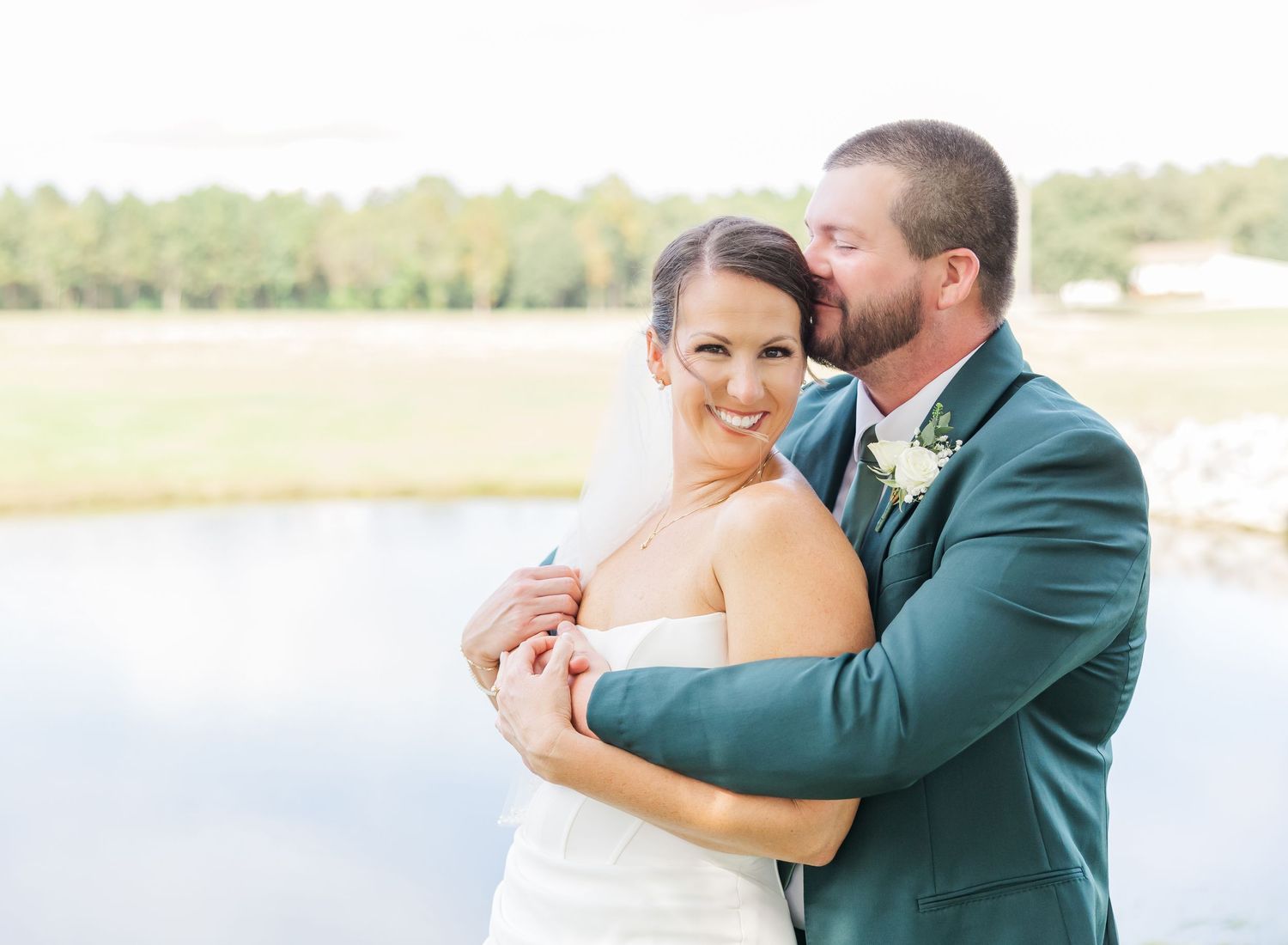 A couple in formal wedding attire embraces by a serene lakeside setting during a romantic outdoor photoshoot.