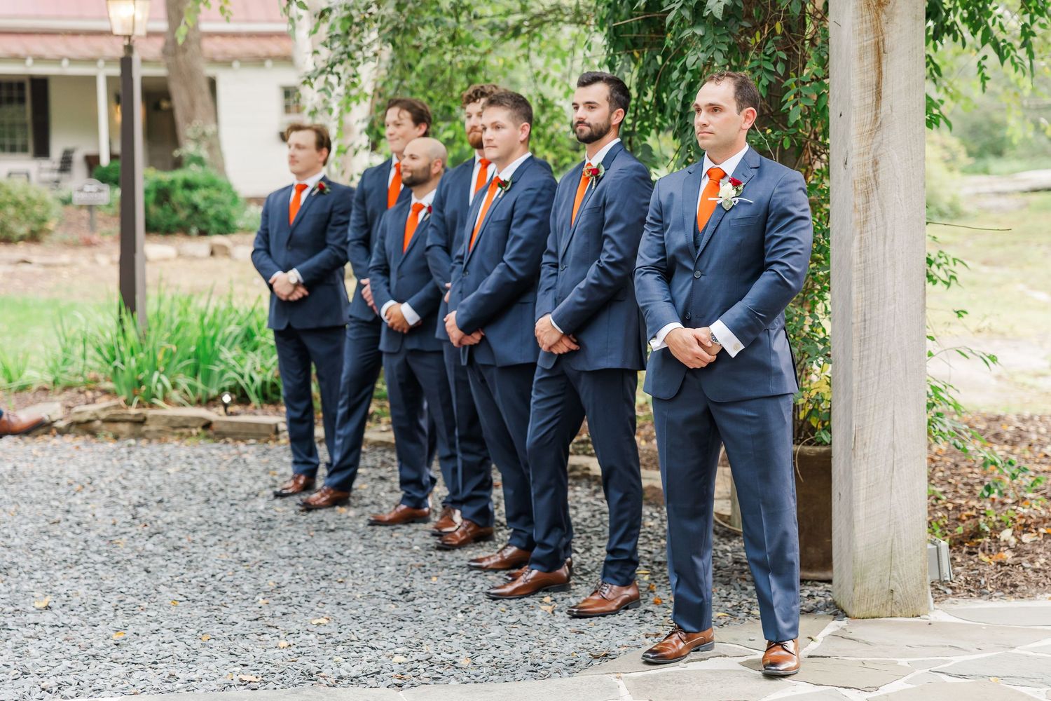 A line of groomsmen in navy suits with orange ties stand on a gravel path next to a wooden post.