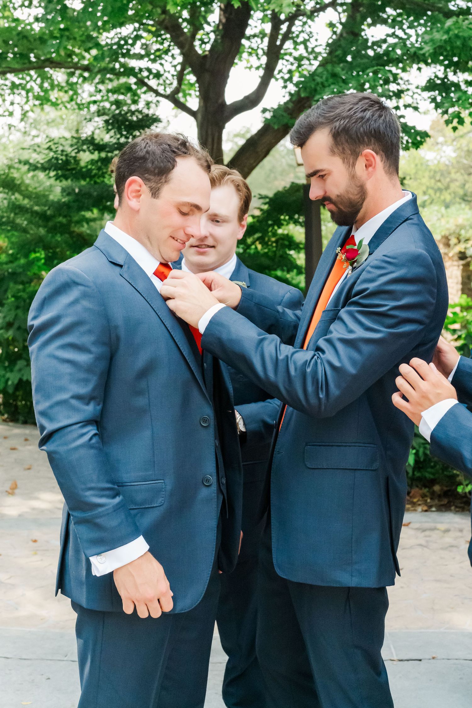 Two groomsmen in navy suits share a lighthearted moment adjusting each other's boutonnieres before a wedding ceremony.