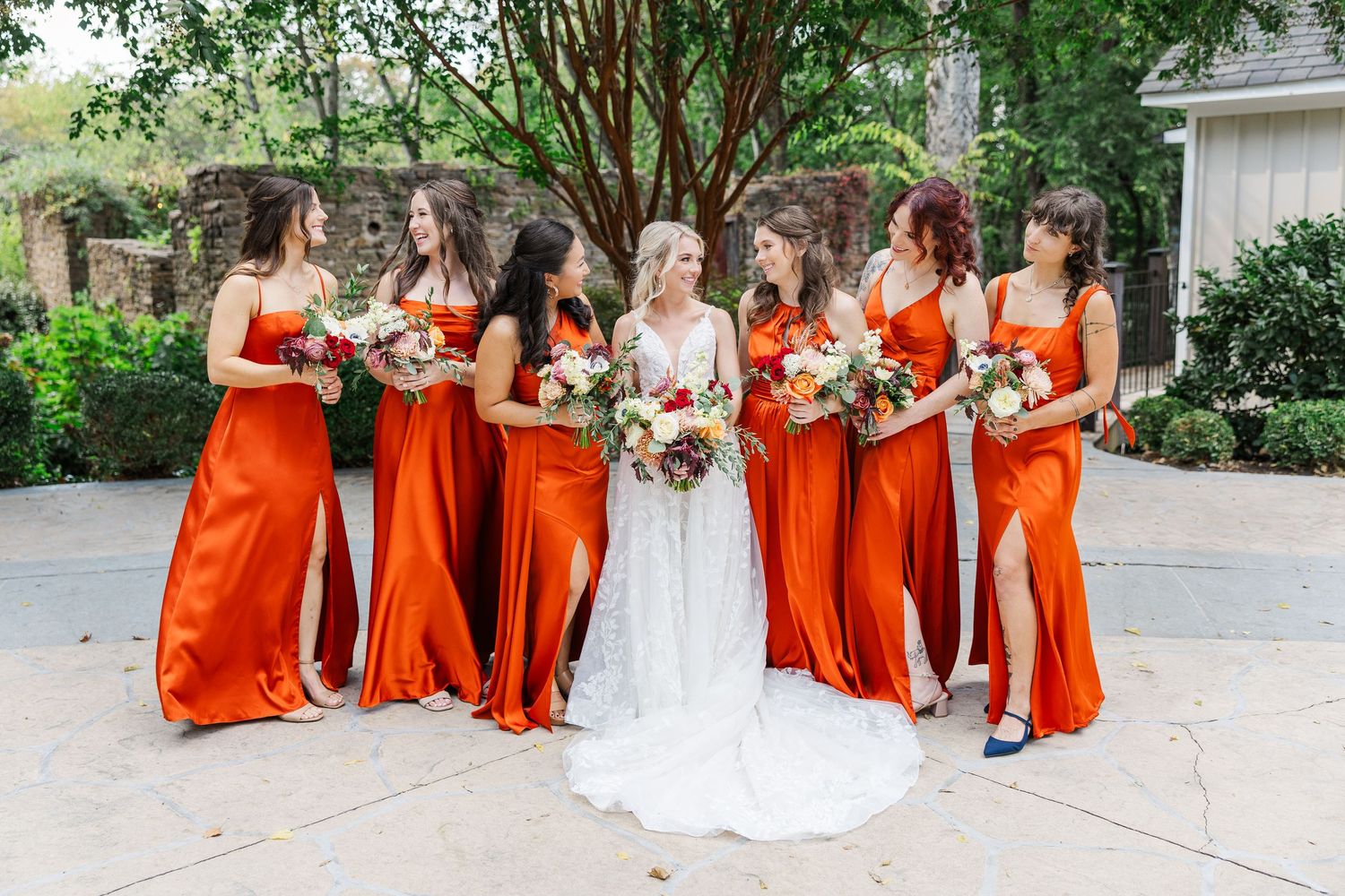 Wedding party wearing bright orange bridesmaid dresses poses together outdoors in a garden setting.