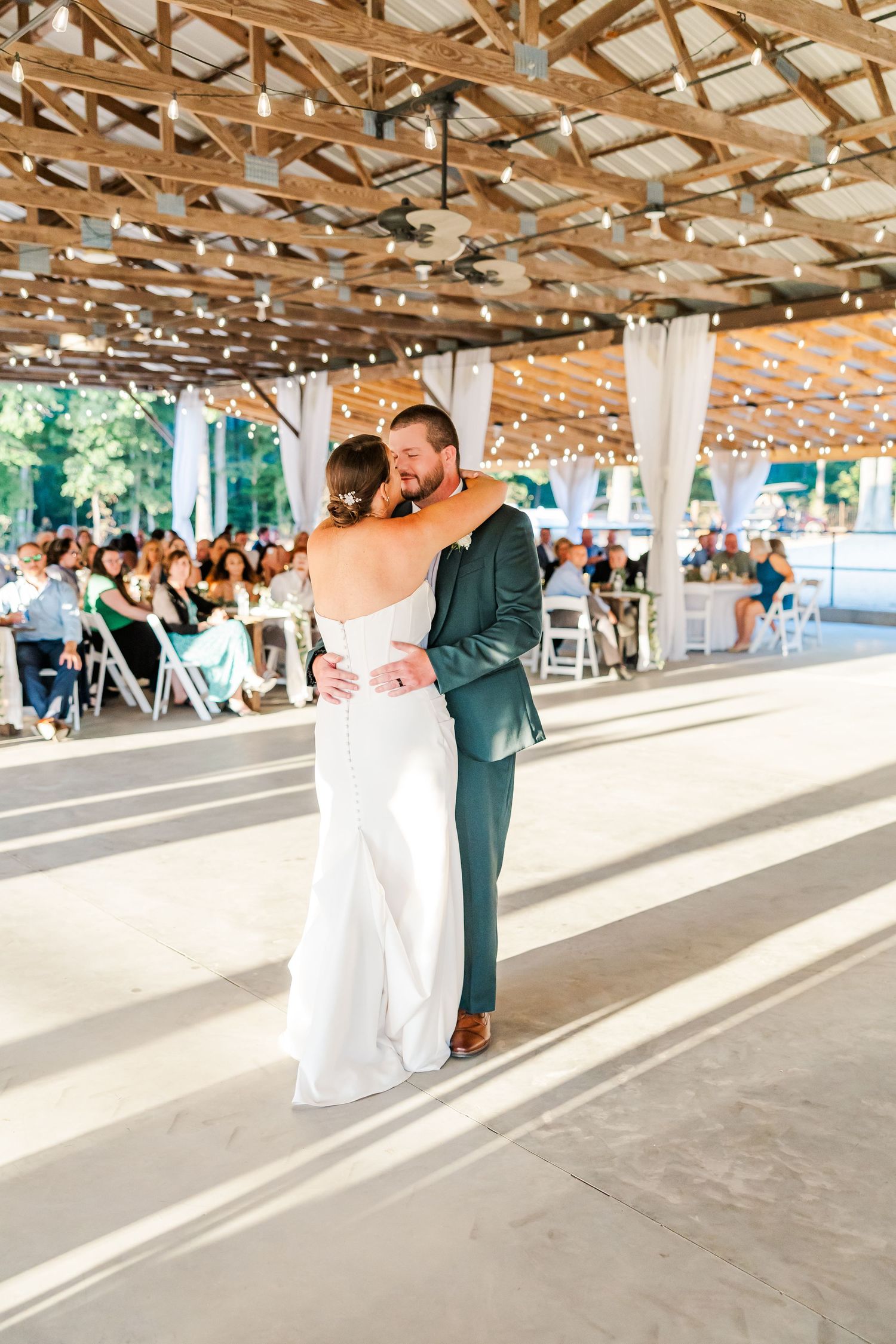 Bride and groom share their first dance under string lights in a rustic open-air wedding venue with guests seated nearby.