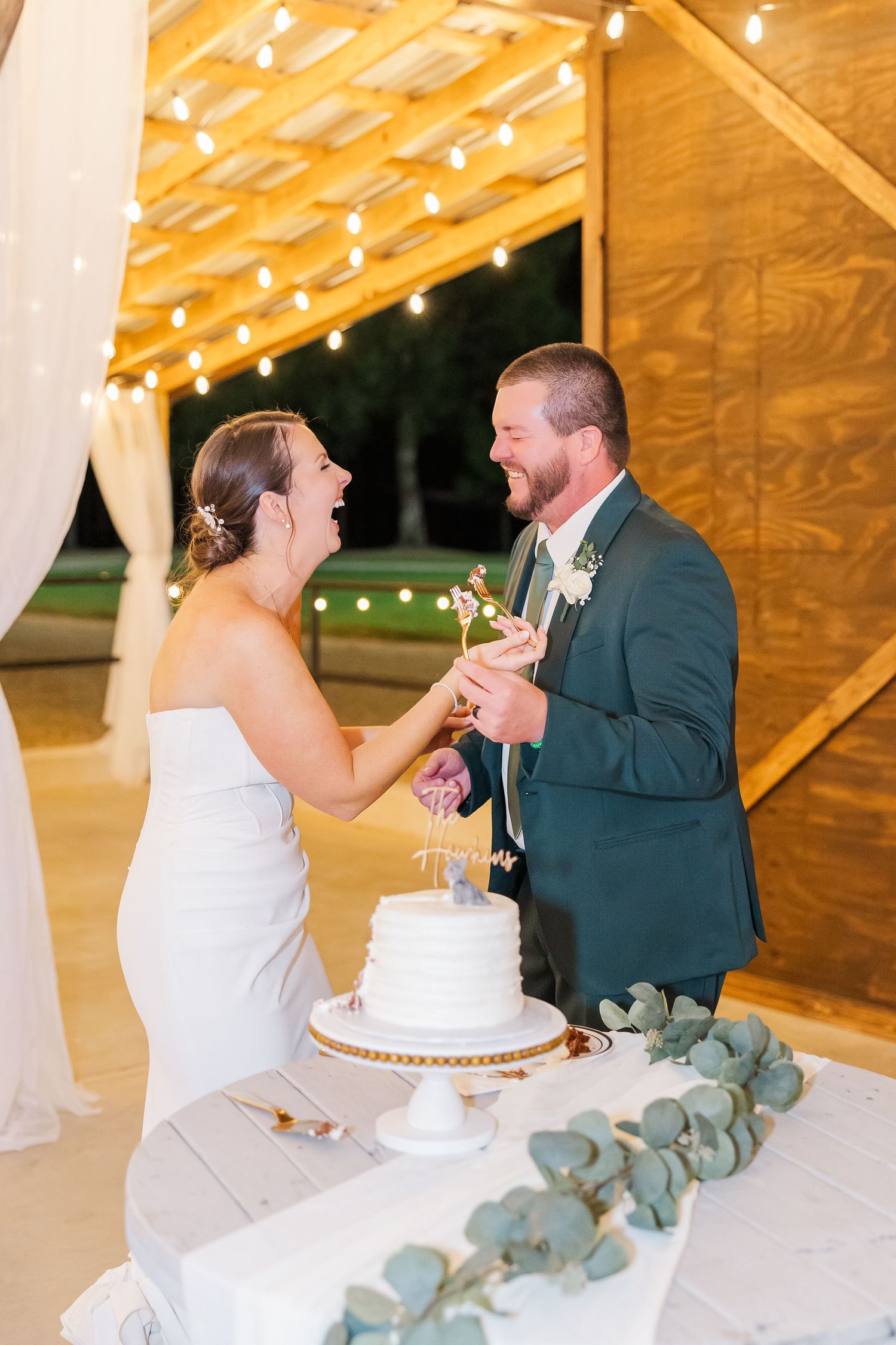 Bride and groom laughing while cutting their white wedding cake at a rustic barn reception with string lights.