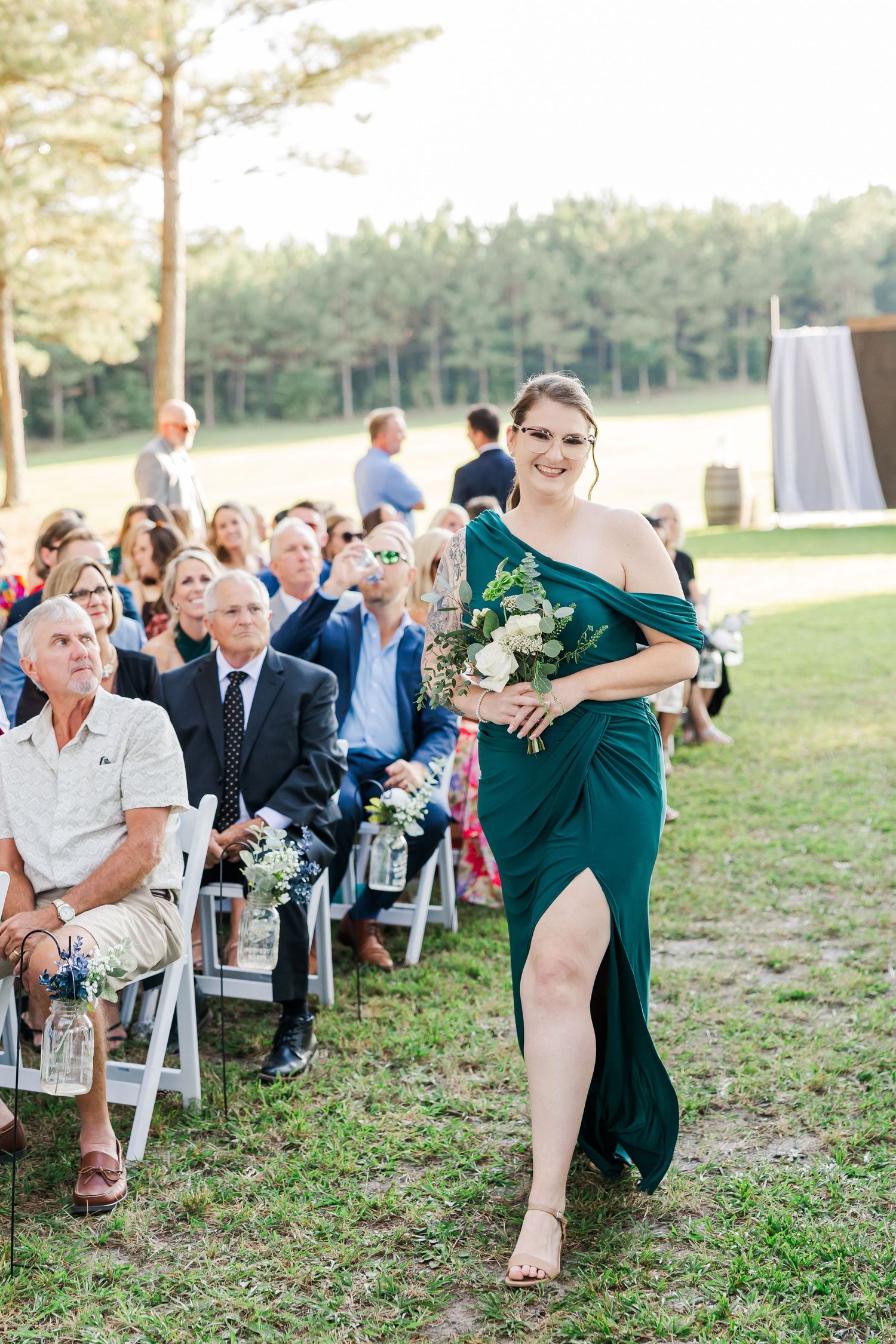 Bridesmaid in emerald green gown walks down outdoor wedding aisle carrying white floral bouquet with guests seated on white chairs.
