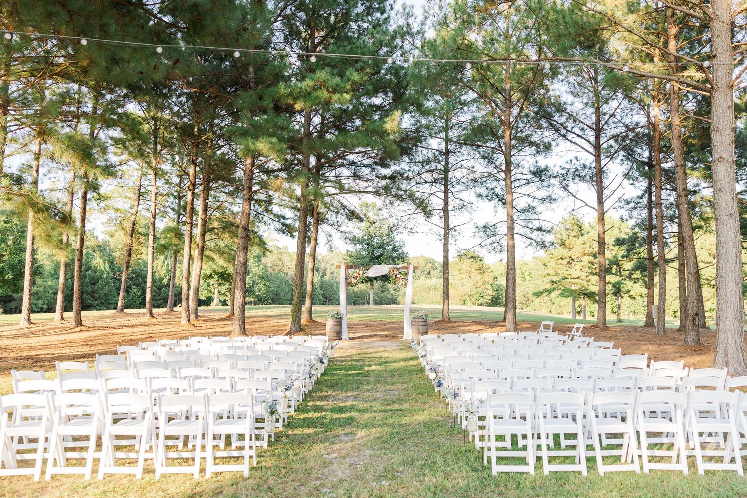 Outdoor wedding ceremony setup with white chairs and wooden arch surrounded by tall pine trees.