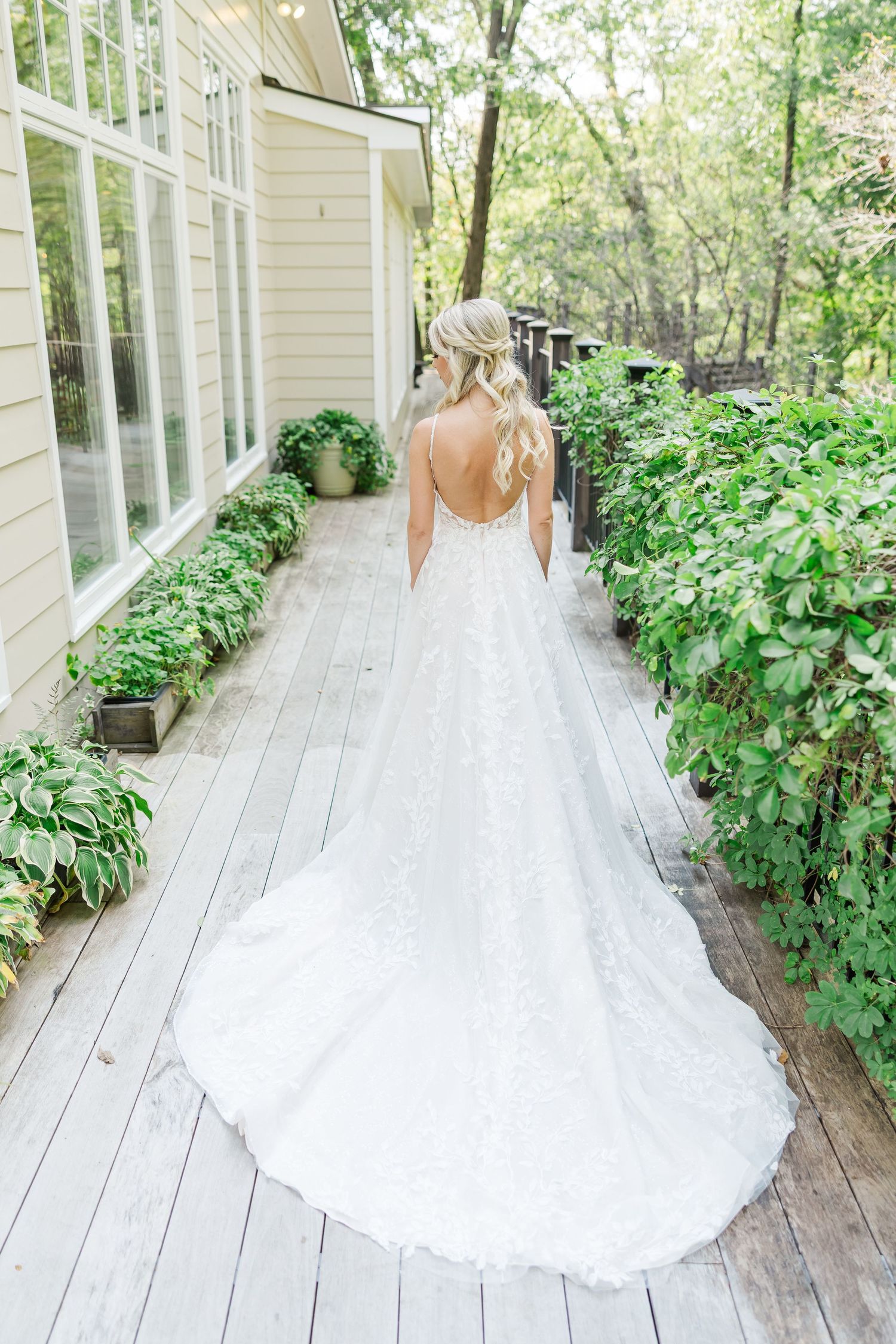 A bride in a flowing white lace gown with train walks along a wooden deck surrounded by lush greenery.
