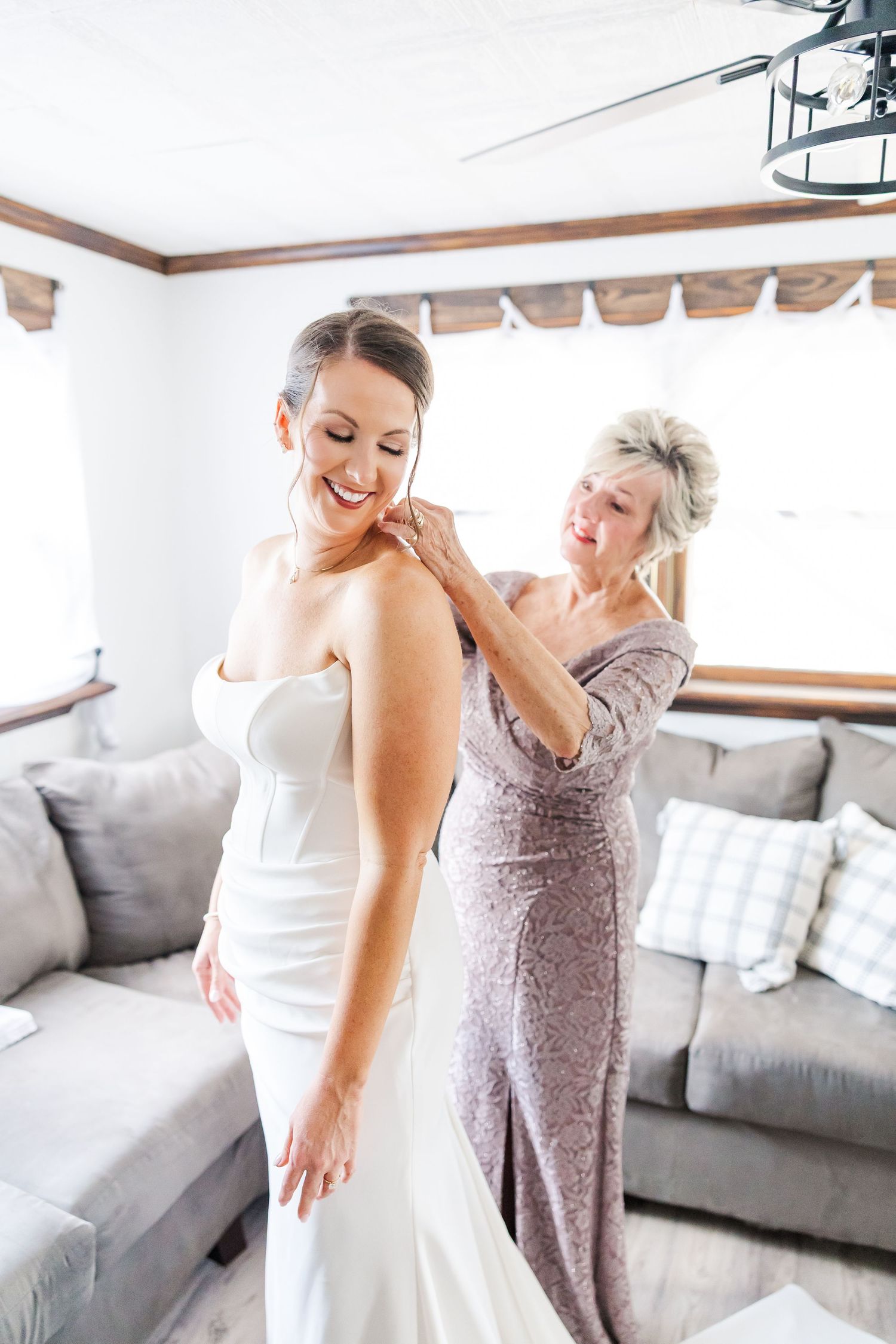Wedding dress preparation scene with two people in a bright room with gray couch and white walls.