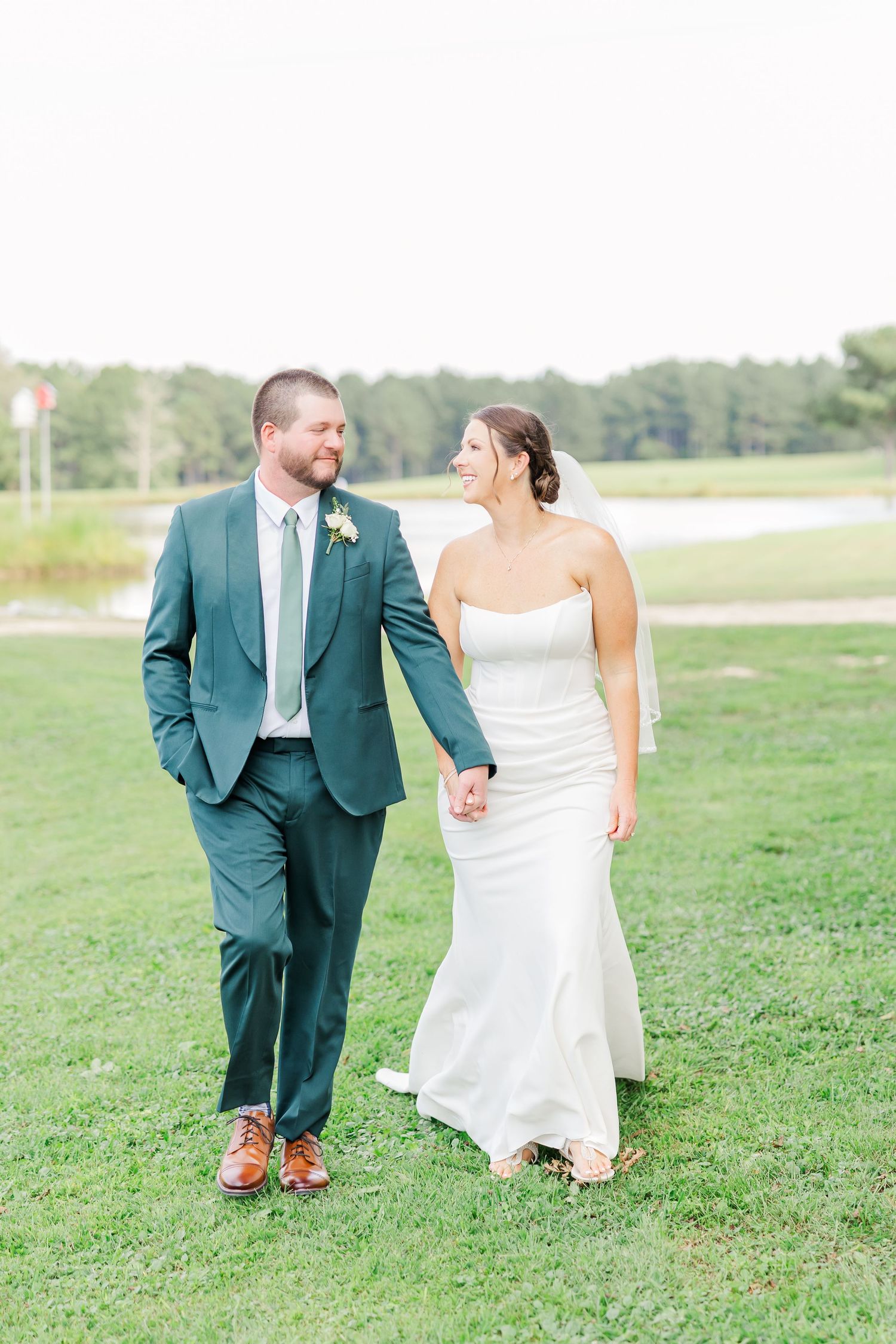 A couple walks hand in hand across a green field during their wedding photoshoot.