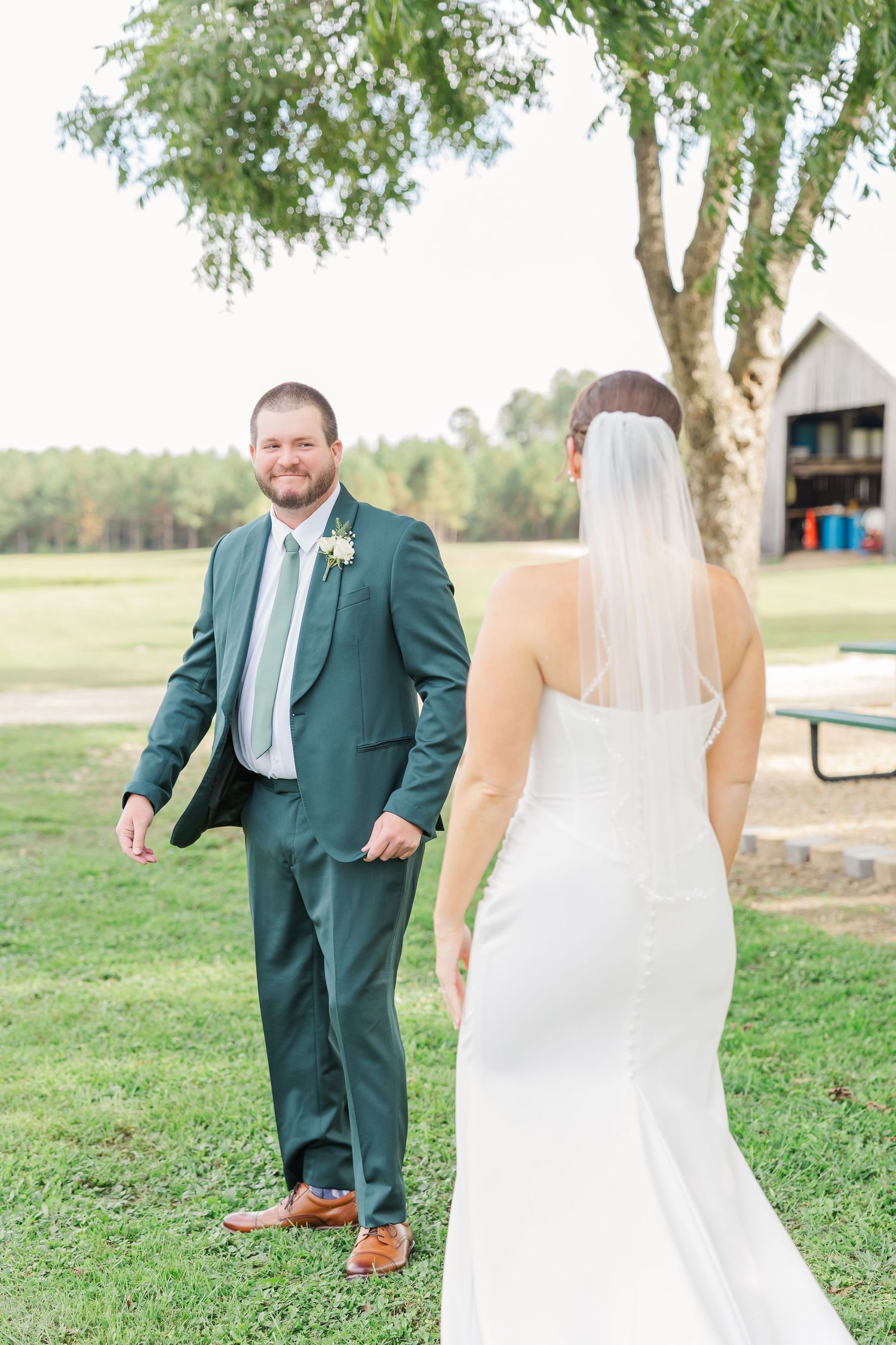 Wedding couple poses outdoors with groom in emerald suit and bride in white dress near trees.