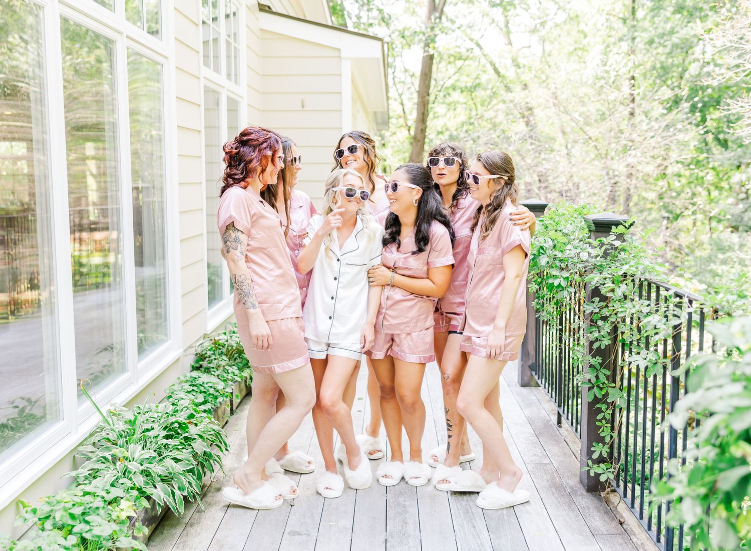 Bridesmaids in matching pink outfits laughing together on a white wraparound porch.