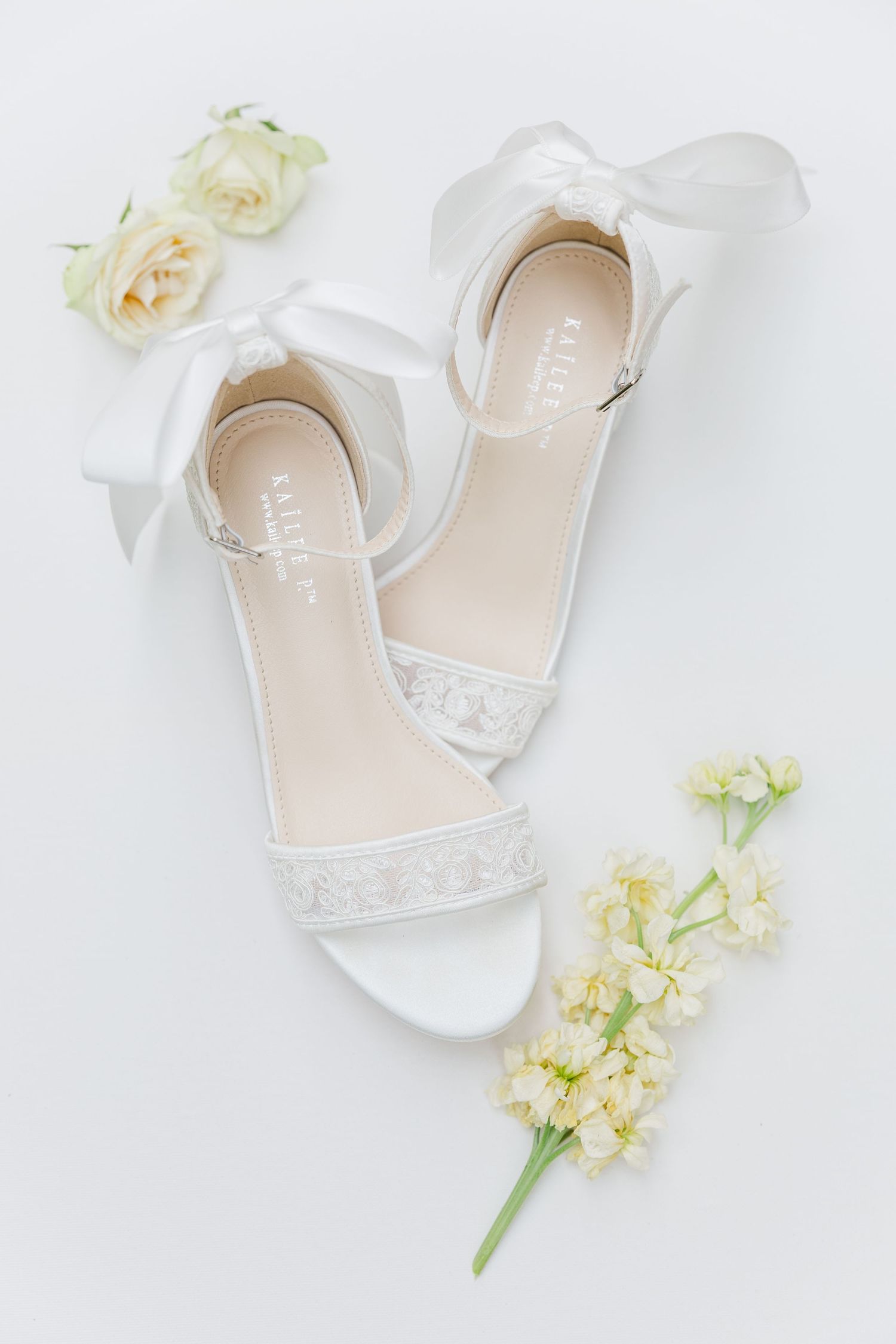 Elegant white bridal shoes with delicate lace details arranged with soft pastel roses and floral accents on light background.