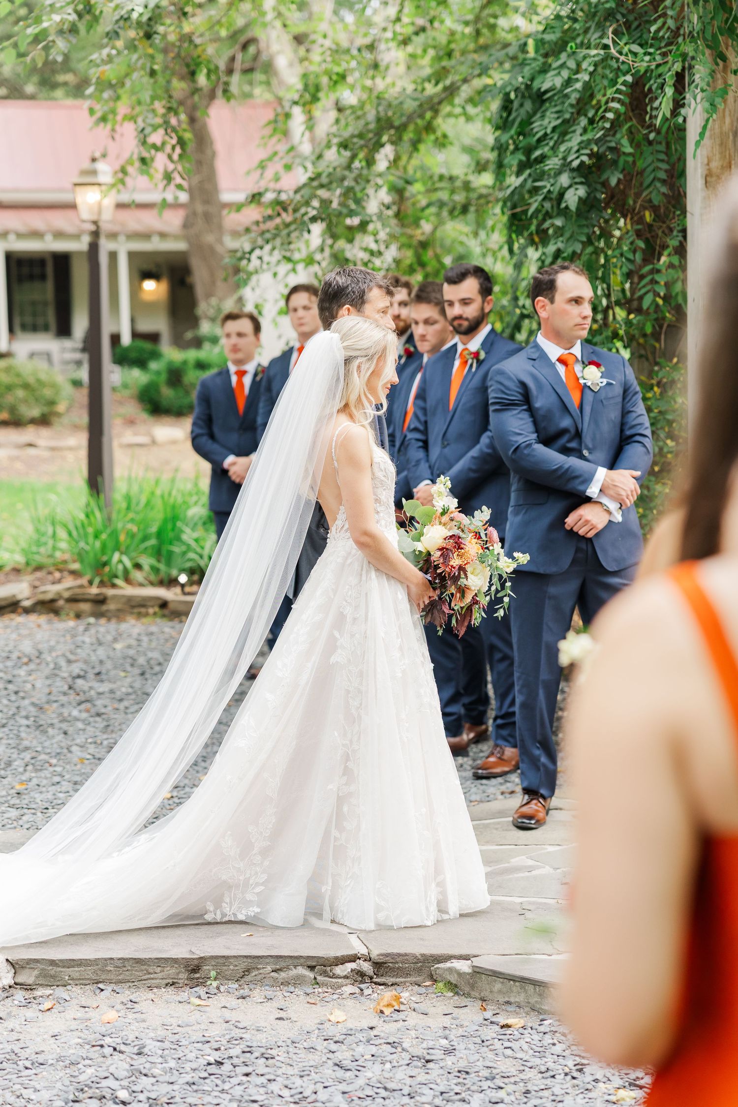 Wedding party gathered on gravel path with bride in flowing white dress and veil alongside navy-suited groomsmen.