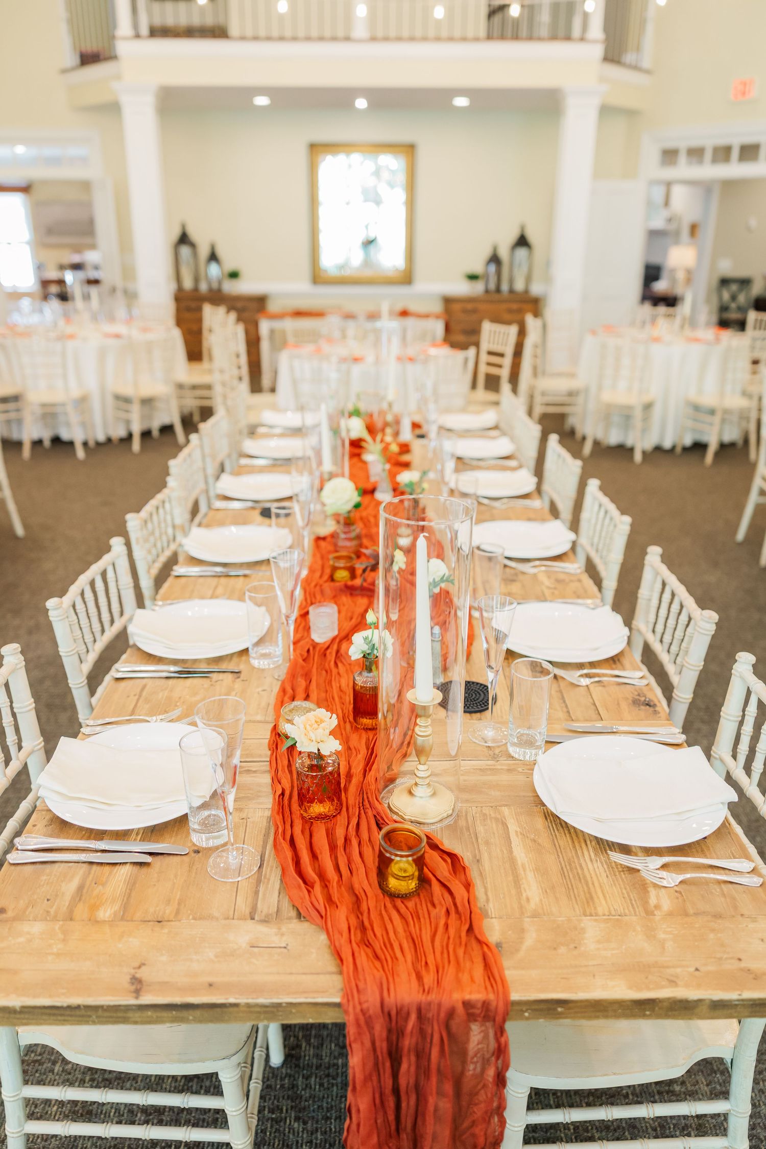Long farmhouse table set for wedding reception with orange runner and white place settings.