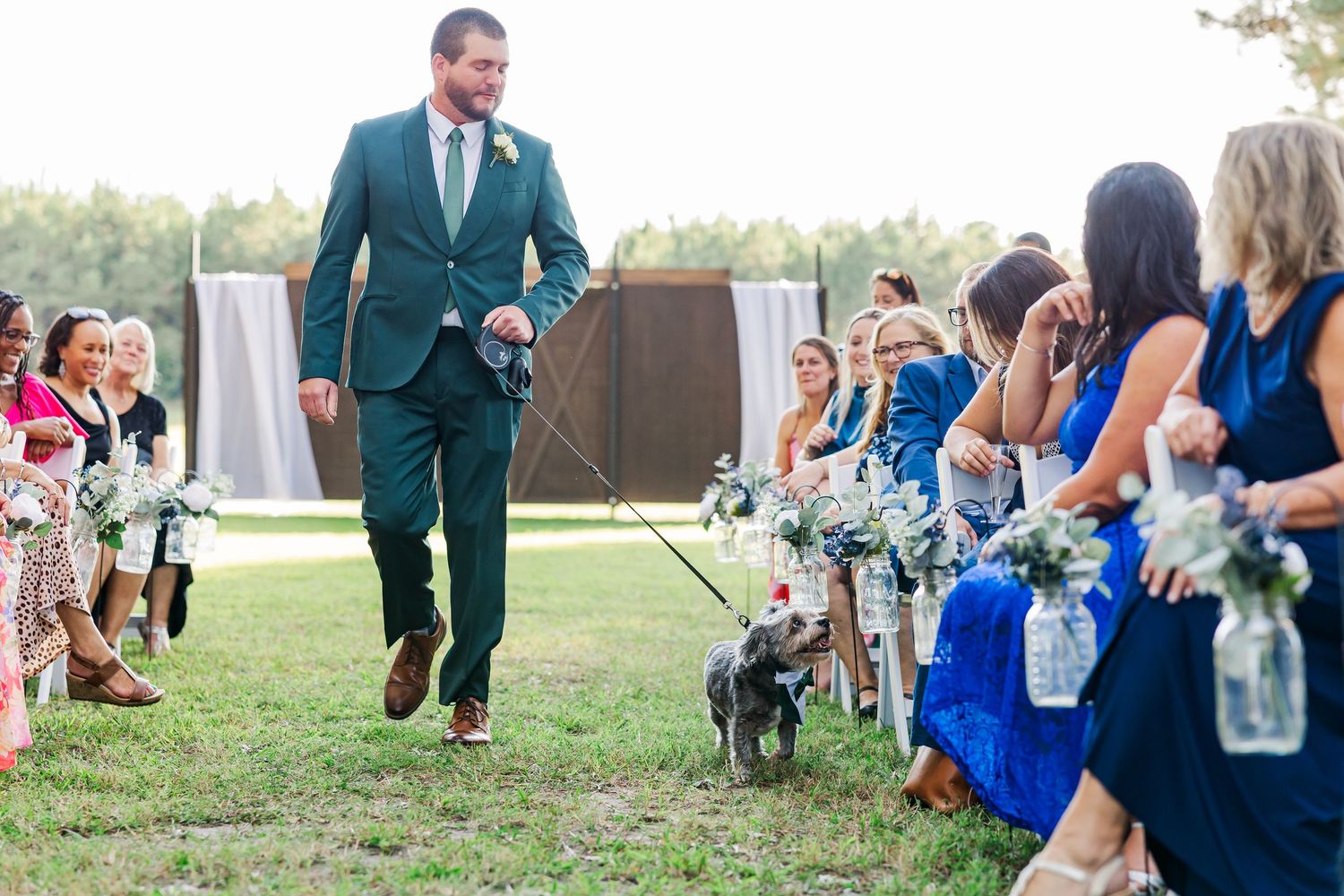 Man in green suit walks dog down wedding aisle outdoors, with guests in blue dresses seated alongside.