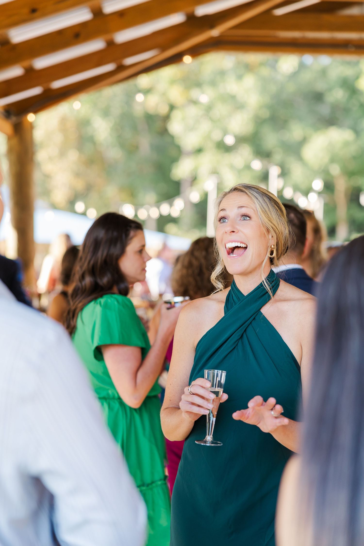 Woman in green halter dress laughing and holding champagne glass at an outdoor wedding reception.