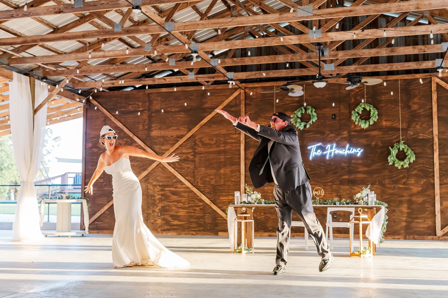 Bride and groom share an energetic first dance in a rustic barn wedding venue decorated with string lights and wreaths.