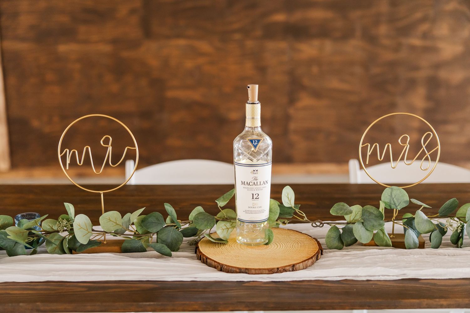 Wedding sweetheart table with Mr and Mrs wire signs flanking a whiskey bottle on wood slice with eucalyptus garland.