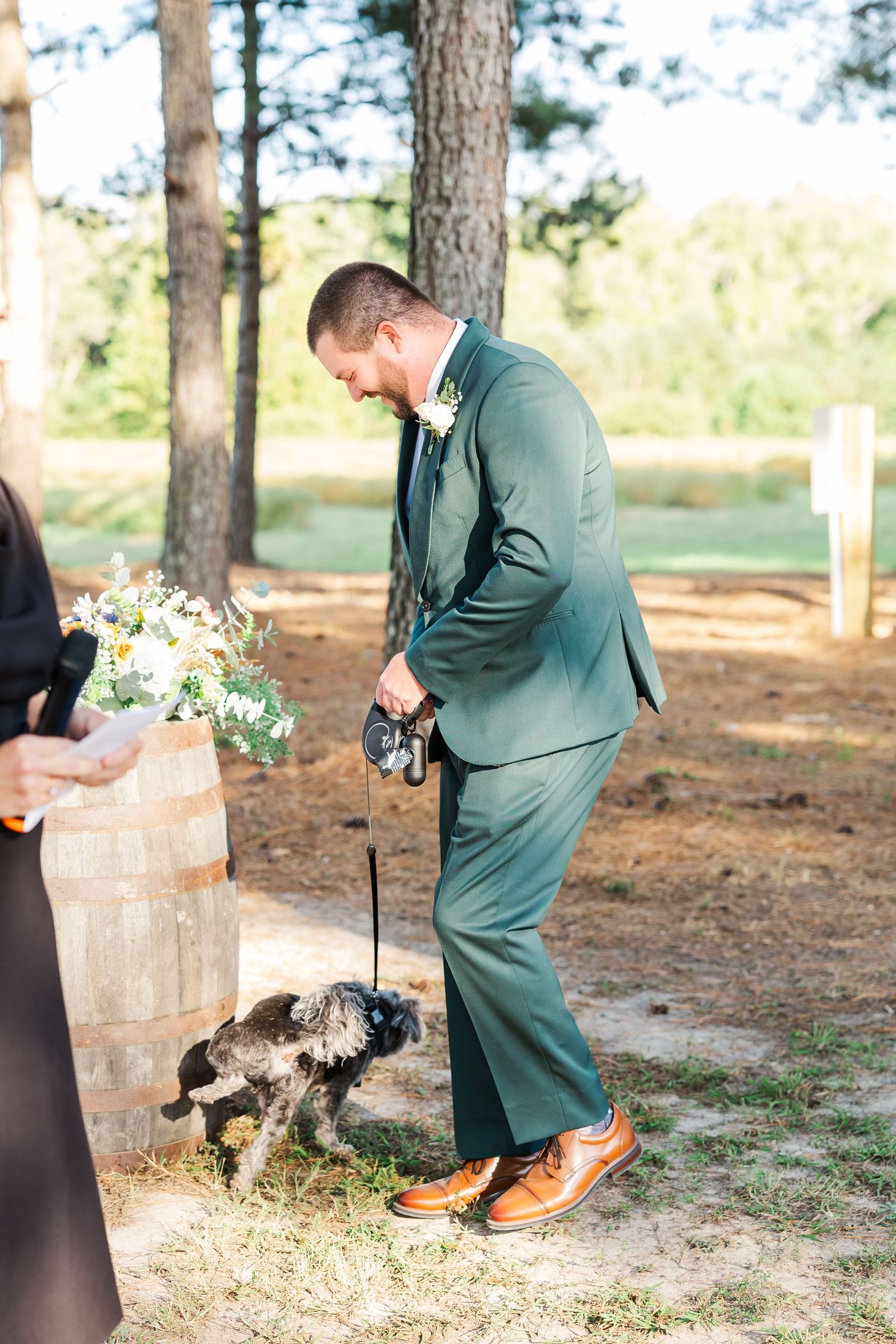 Groom in green suit with boutonniere bends to greet small dog on leash at outdoor wedding ceremony.