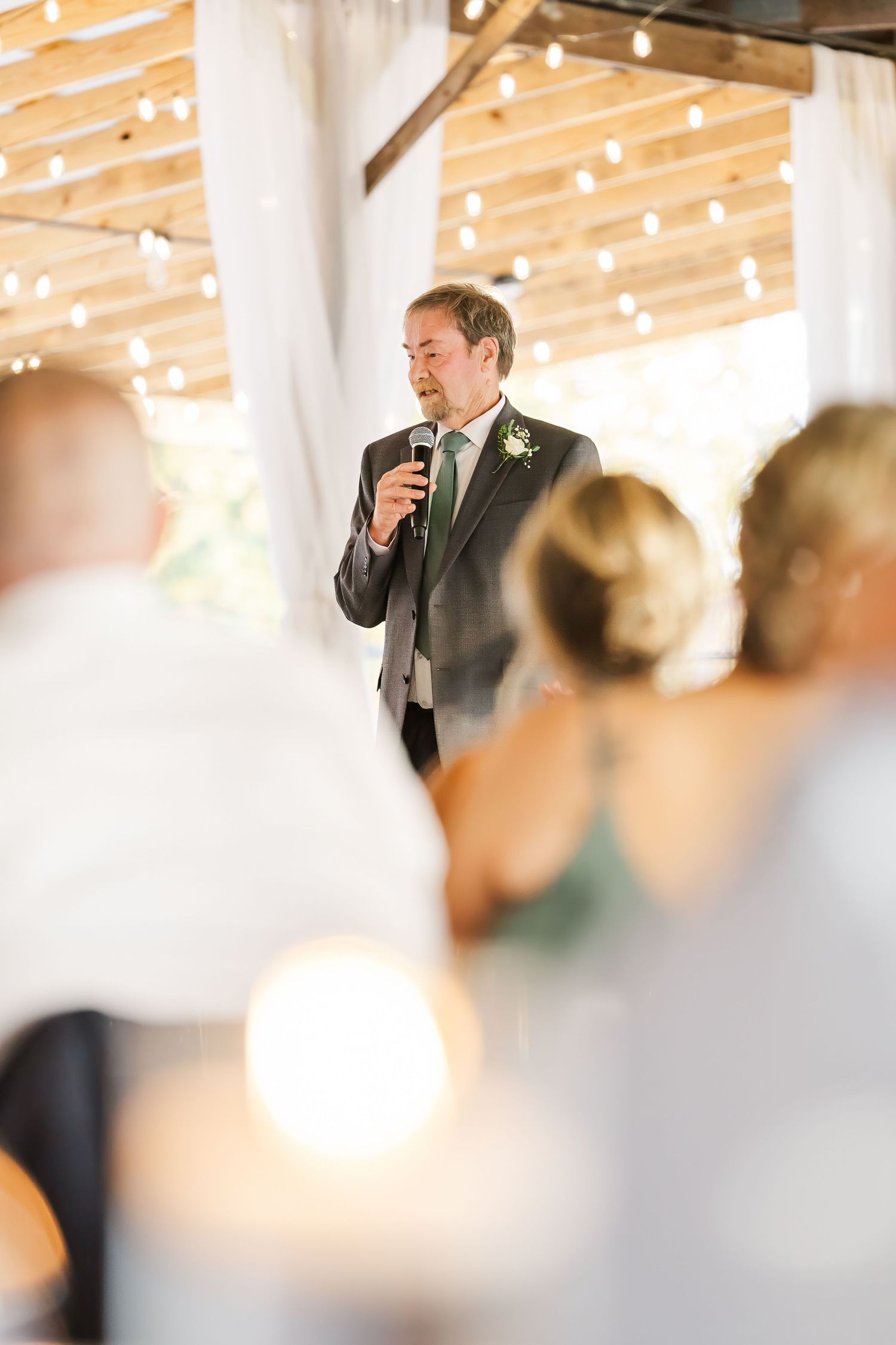 Groom delivering a heartfelt speech at wedding reception under a rustic wooden ceiling with warm string lights.