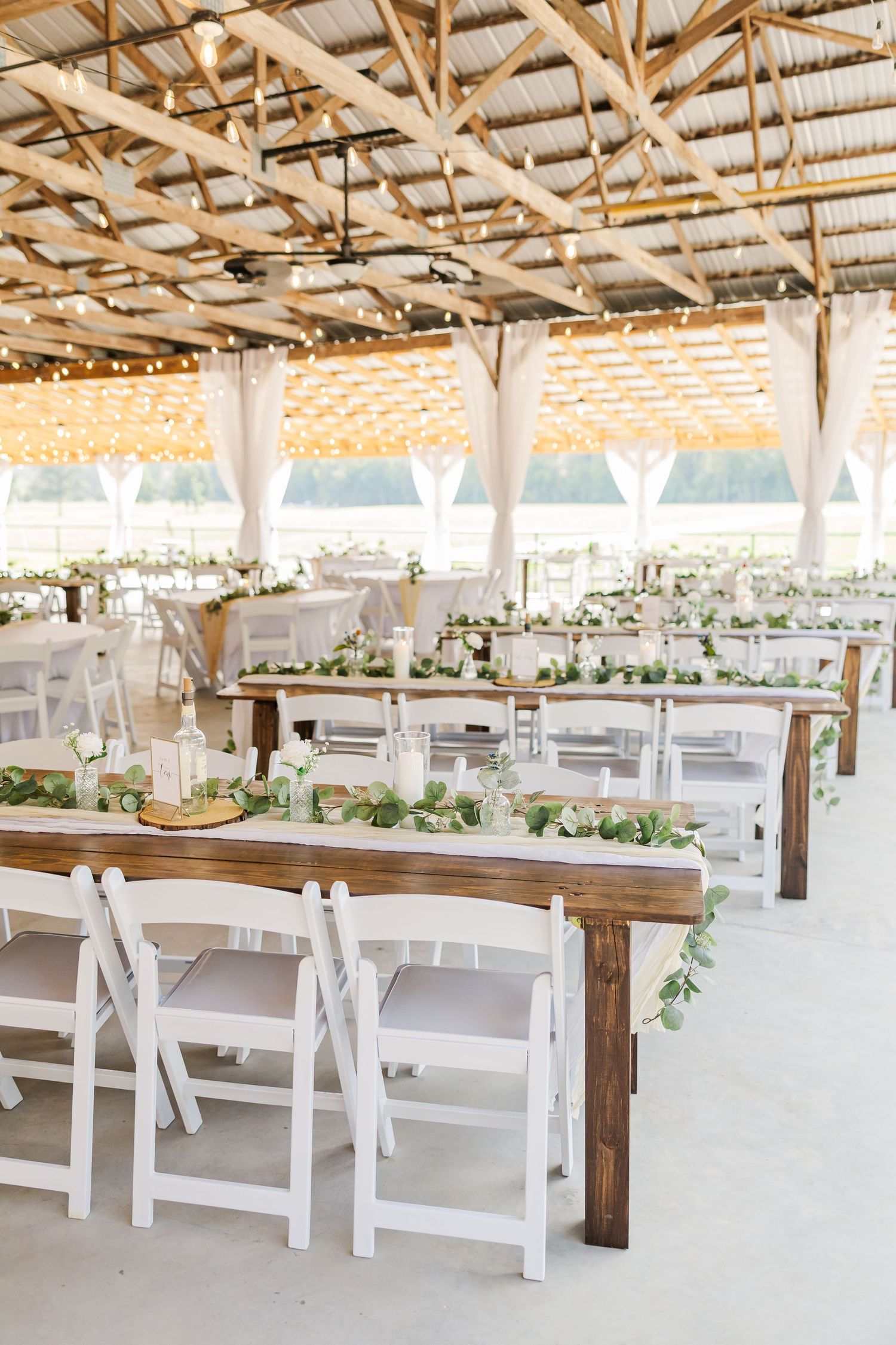 Rustic barn wedding reception with wooden farm tables, white folding chairs, greenery garland runners, and string lights overhead.