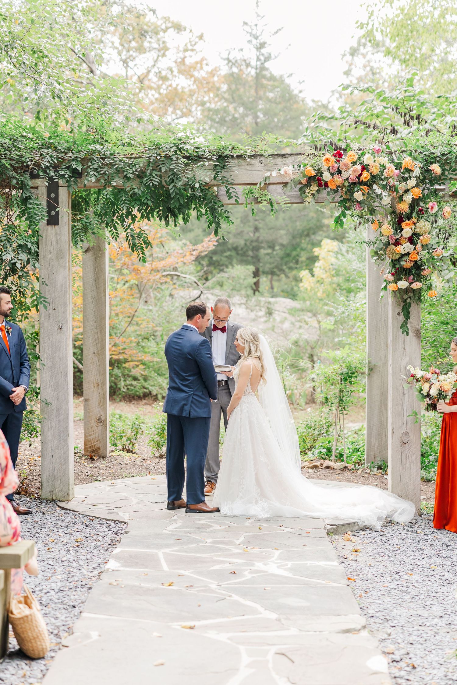 A romantic outdoor wedding ceremony takes place under a floral-adorned pergola on a gravel path in a garden setting.