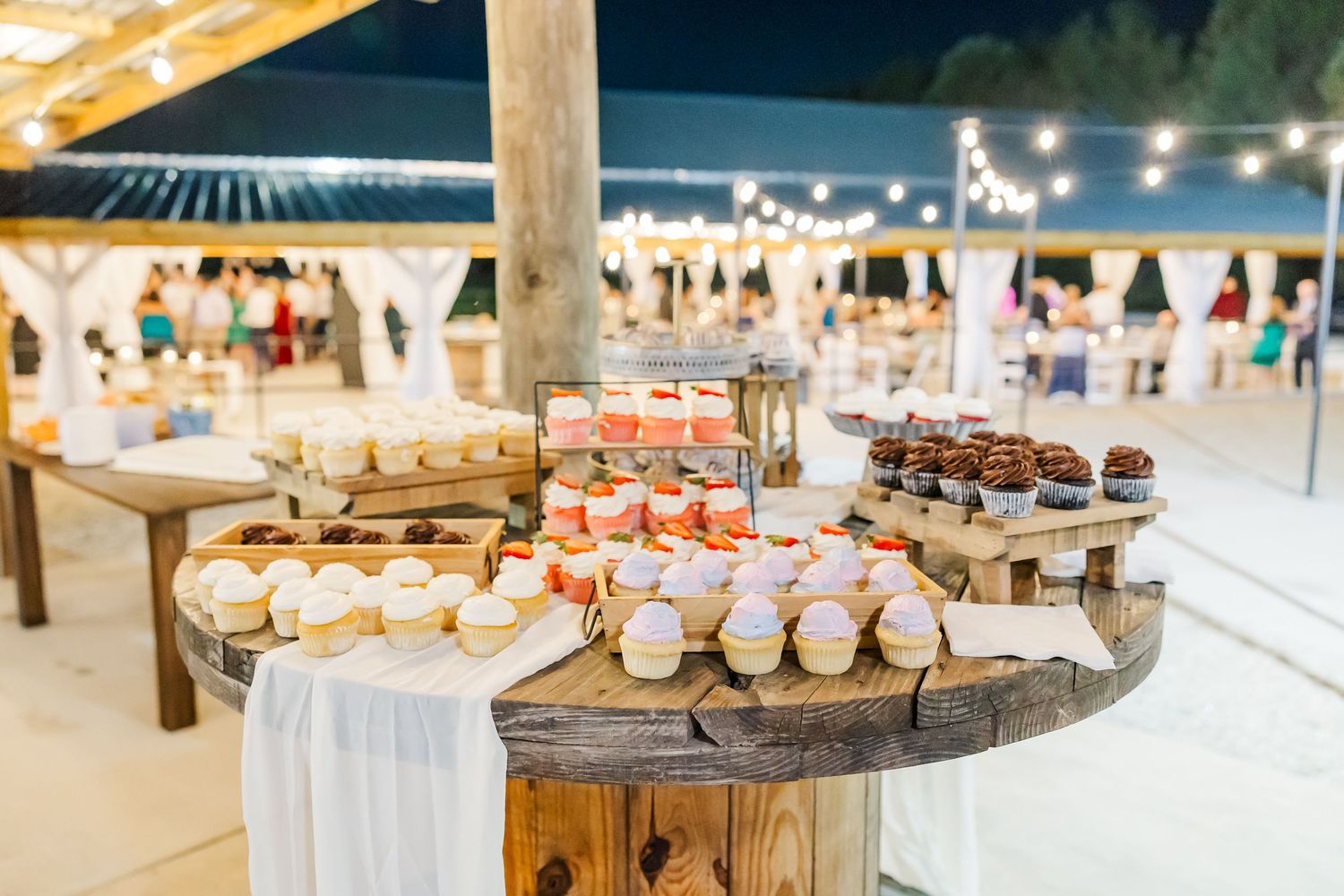 Rustic wooden spool dessert table with cupcakes and treats at an outdoor wedding reception.