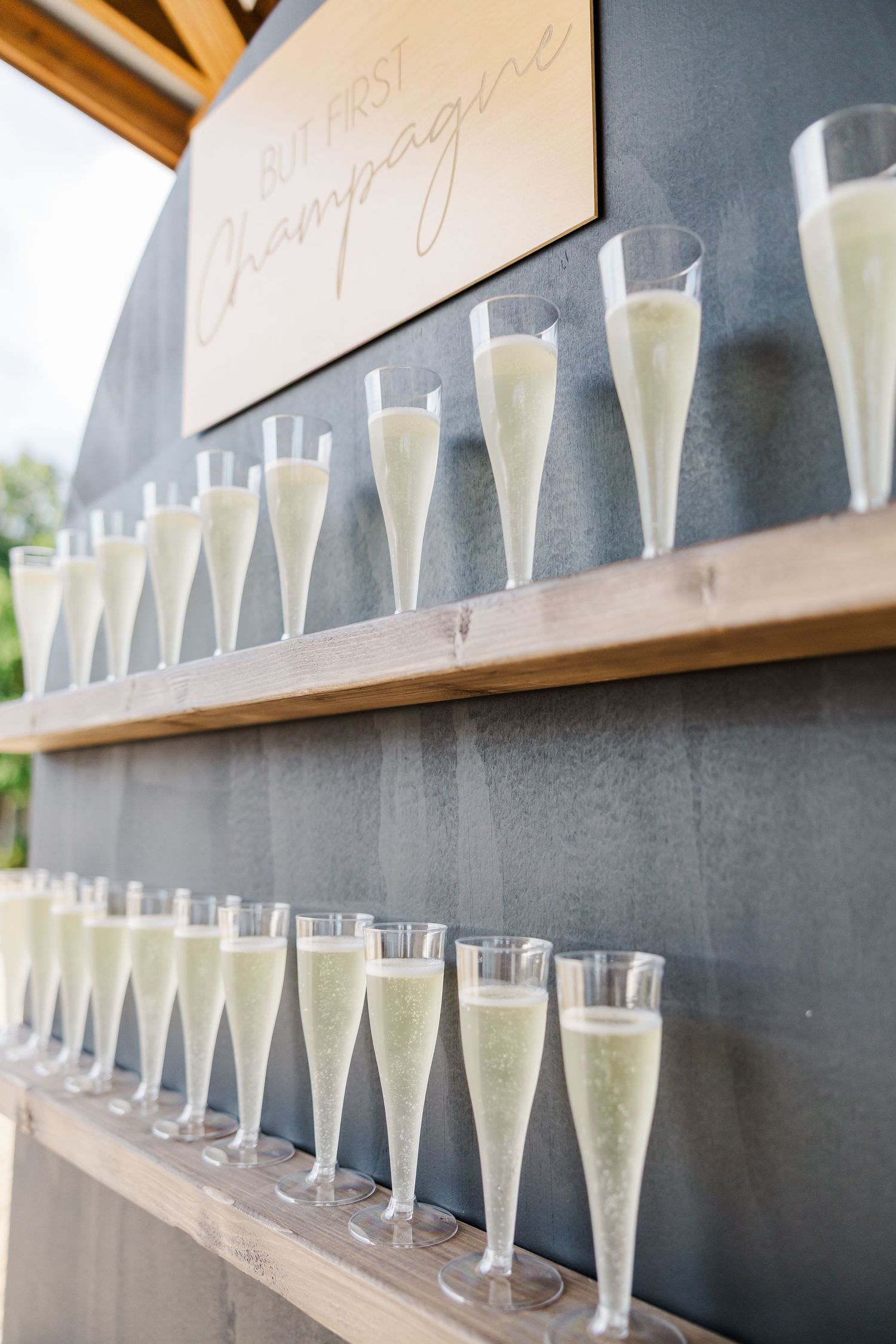 Rows of champagne flutes on wooden shelves with a But First Champagne sign at a wedding reception.