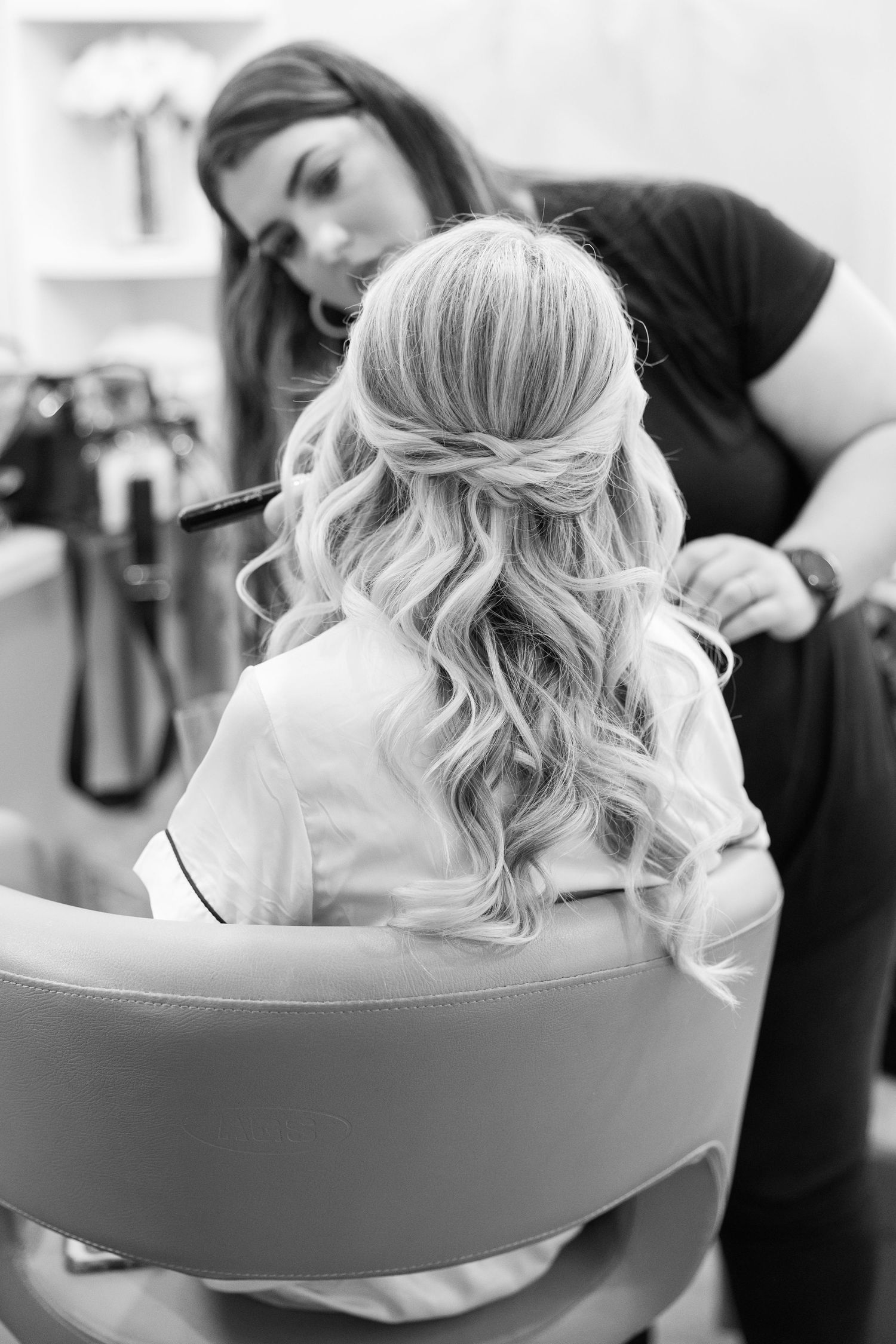 A hairstylist works on styling a blonde updo with soft waves and curls in a salon chair.