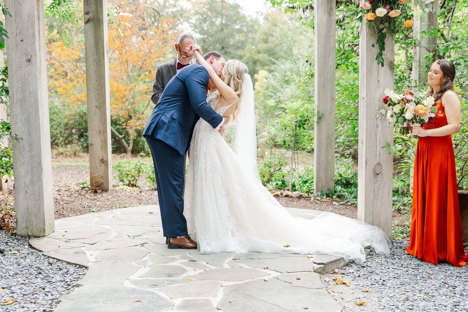 A couple shares a romantic kiss under an elegant outdoor pergola while a bridesmaid in red looks on during the ceremony.