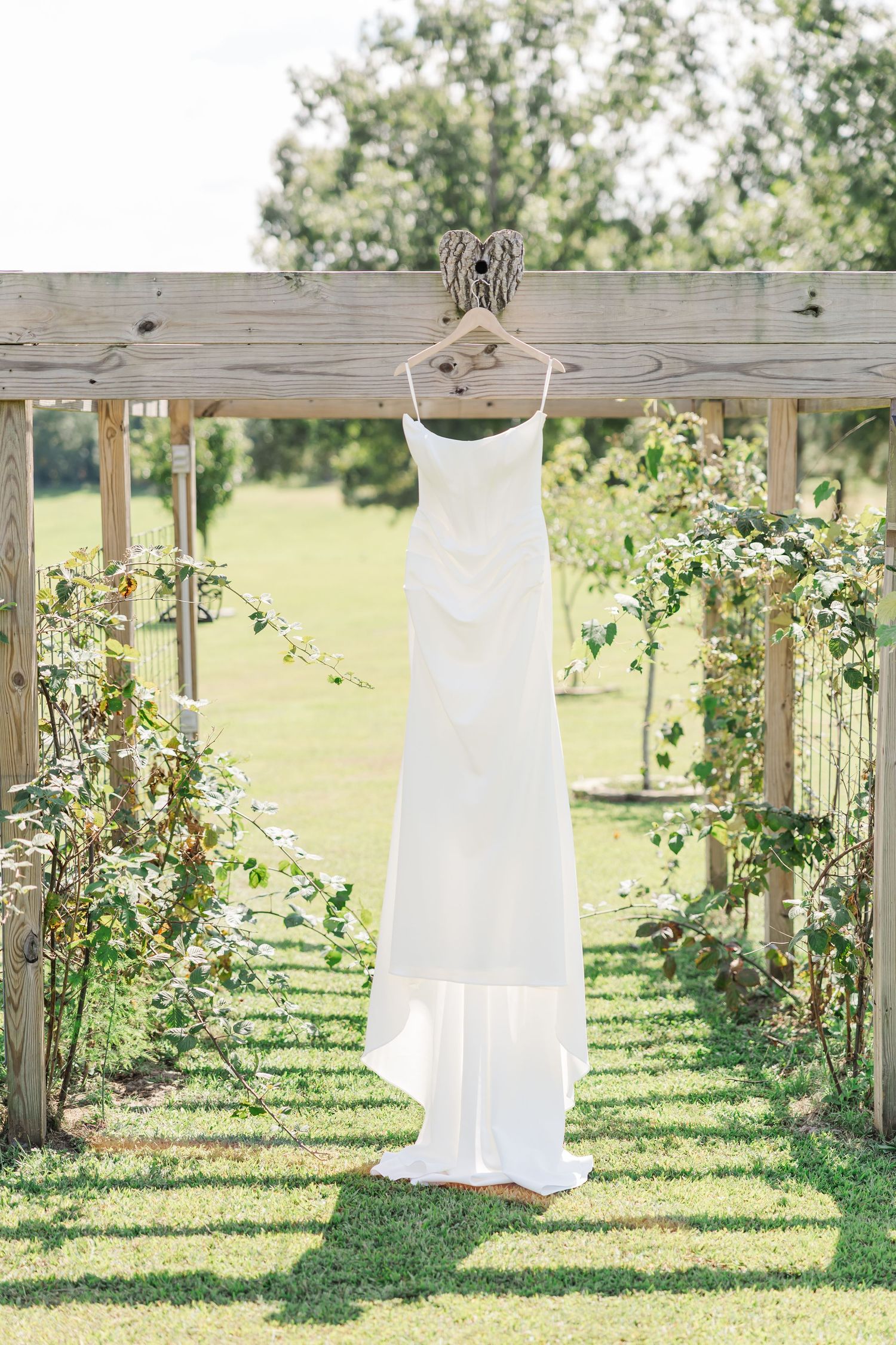 A white wedding dress hangs on a rustic wooden pergola in a garden setting with blooming roses and greenery.