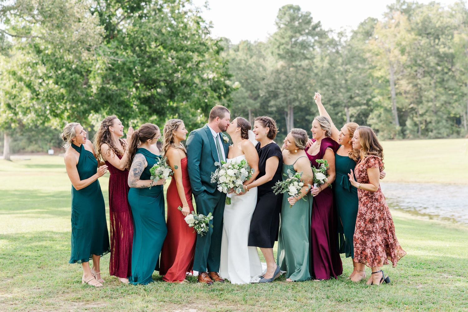 Large wedding party group poses together outdoors wearing jewel-toned dresses and formal attire.