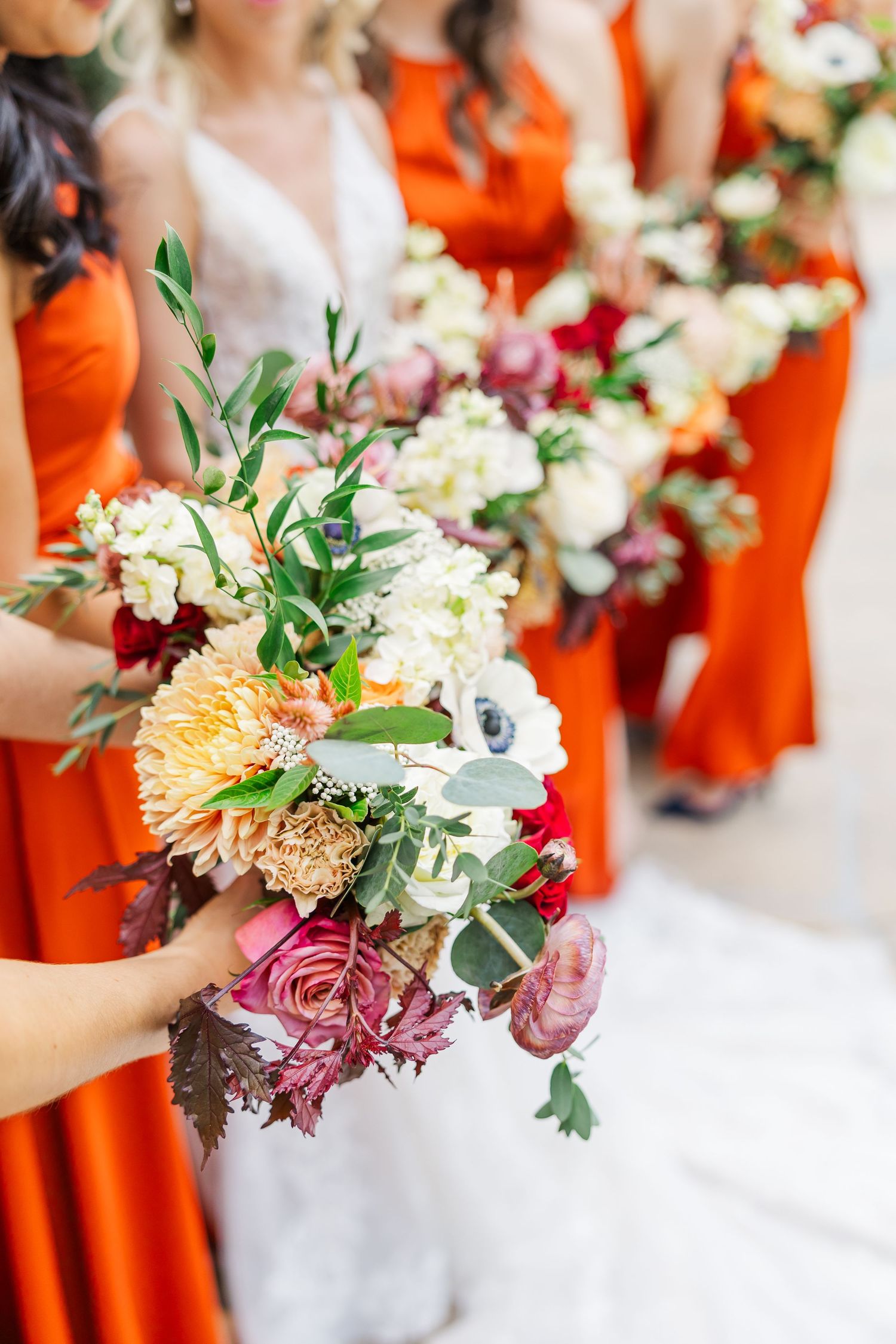 Bridesmaids in vibrant orange dresses hold elegant bouquets with peach, burgundy and white flowers against a light background.