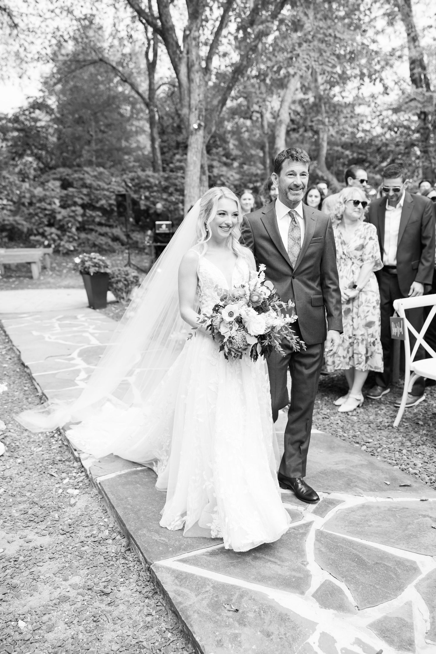 A wedding processional on a stone path with a bride in white gown carrying a floral bouquet.
