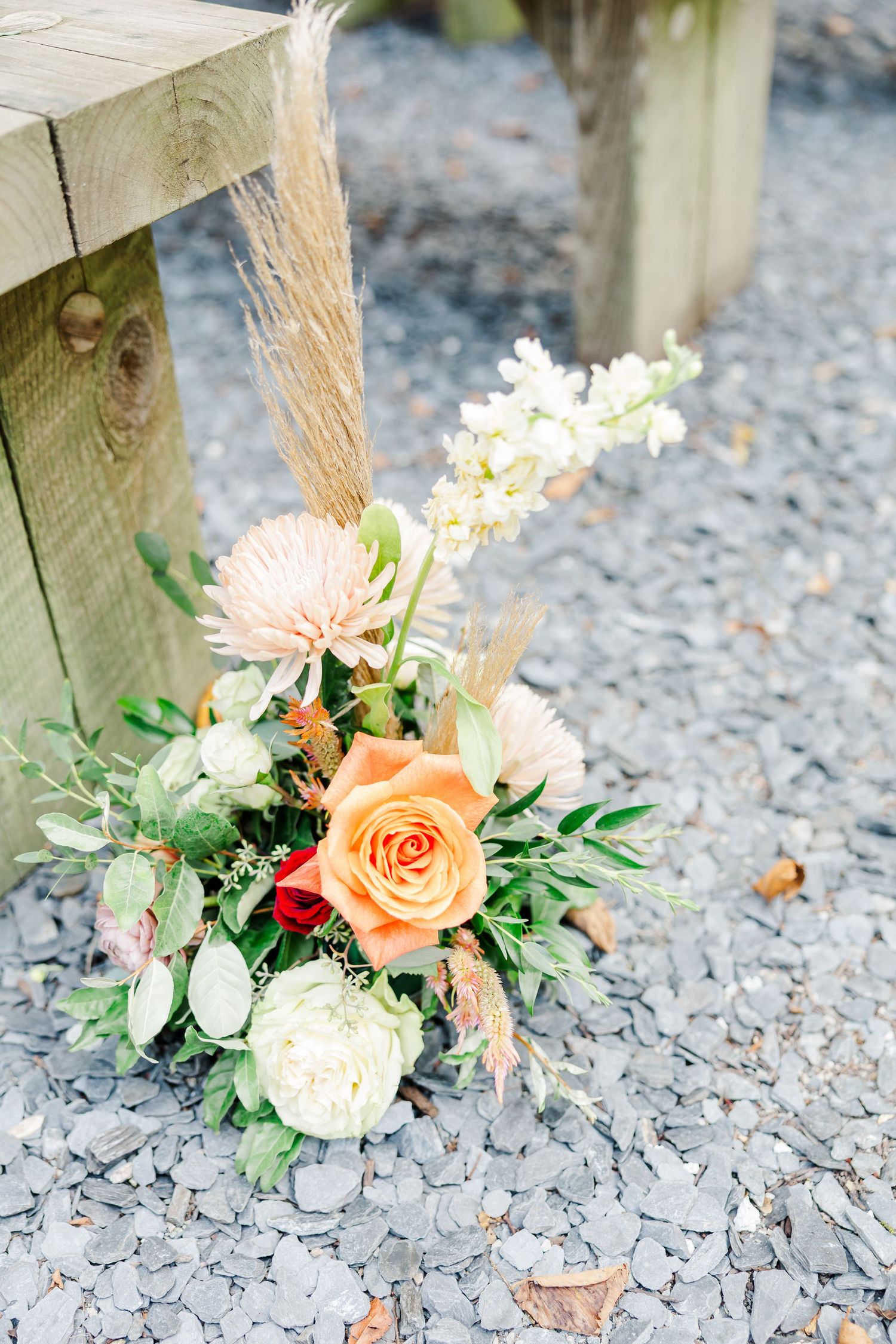 Small floral arrangements with peach roses and white flowers sit at the base of wooden posts on gravel ground.