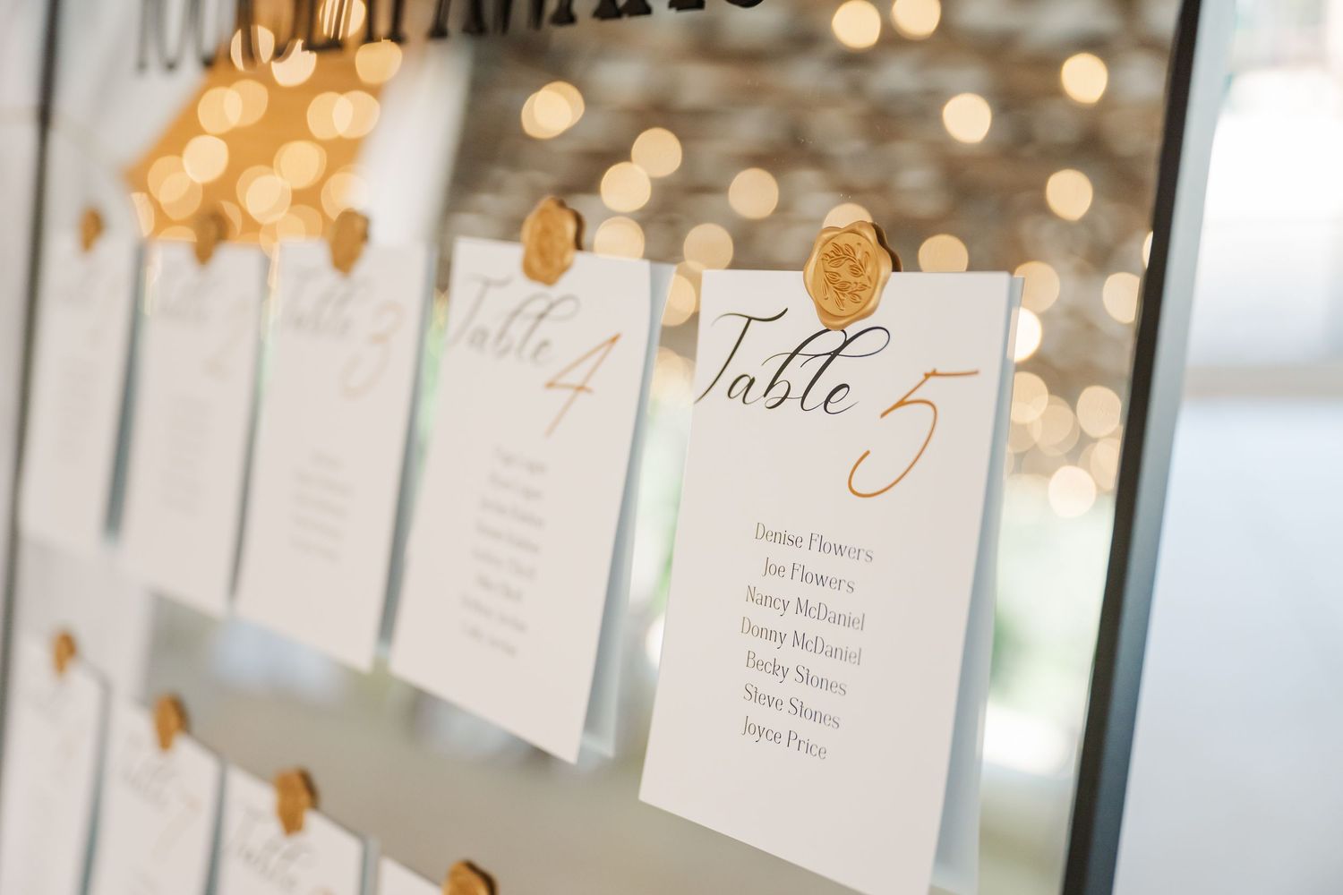 Elegant wedding seating chart display with white cards and gold wax seals on a mirror, featuring Table 5 in script font.