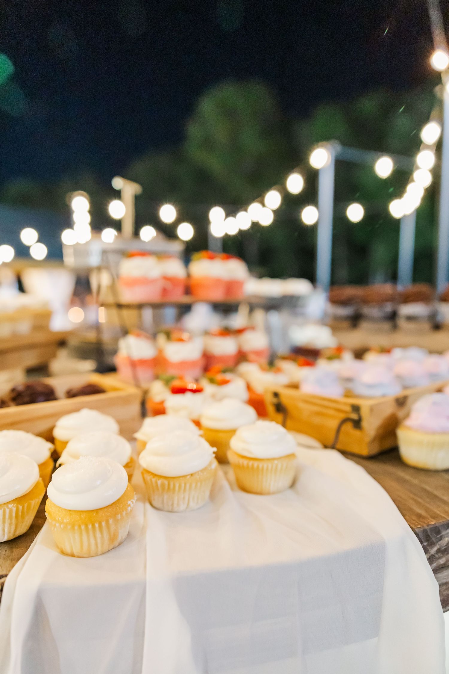 Elegant wedding dessert table with white frosted cupcakes and assorted sweets under string lights.