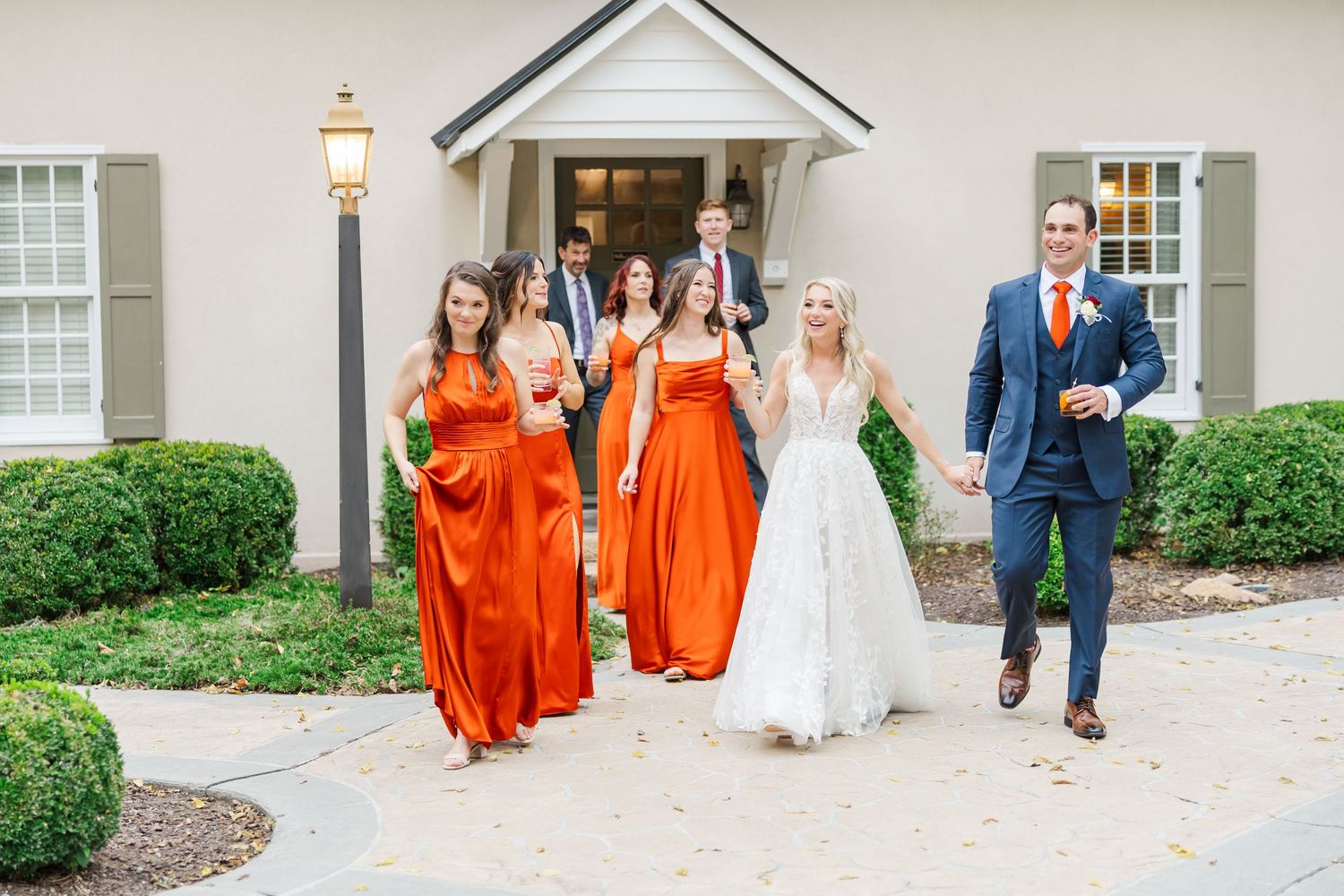 Wedding party in orange bridesmaid dresses walking together on a stone path outside a traditional building.