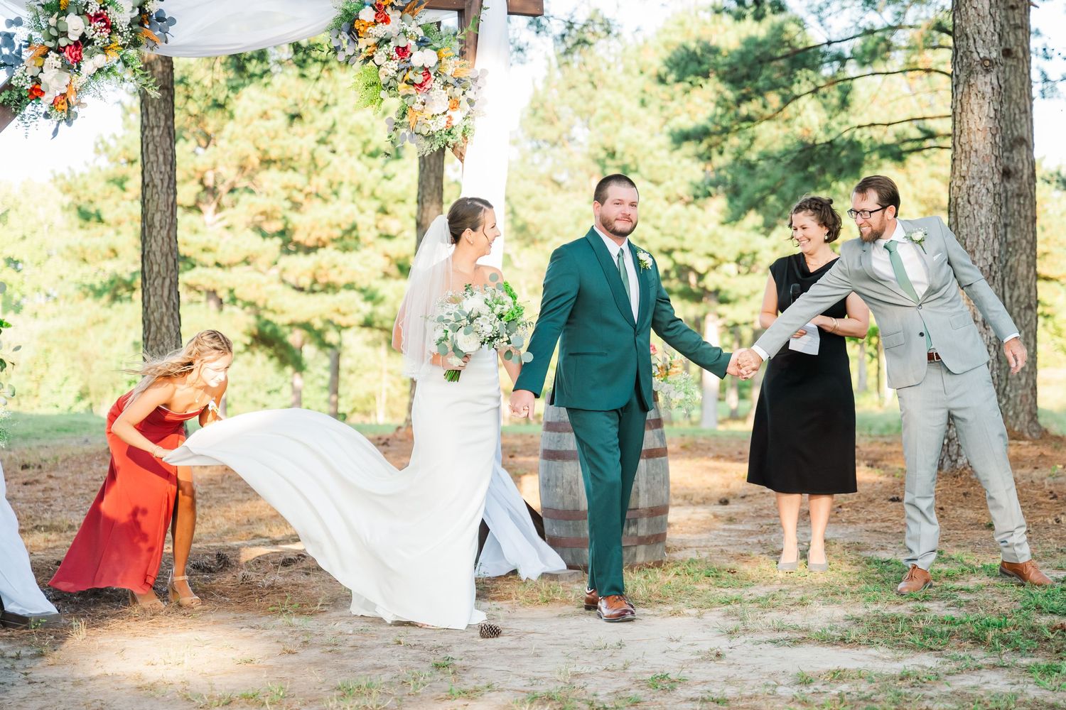 Bride and groom walk joyfully after outdoor wedding ceremony under floral arch, surrounded by guests in colorful attire.