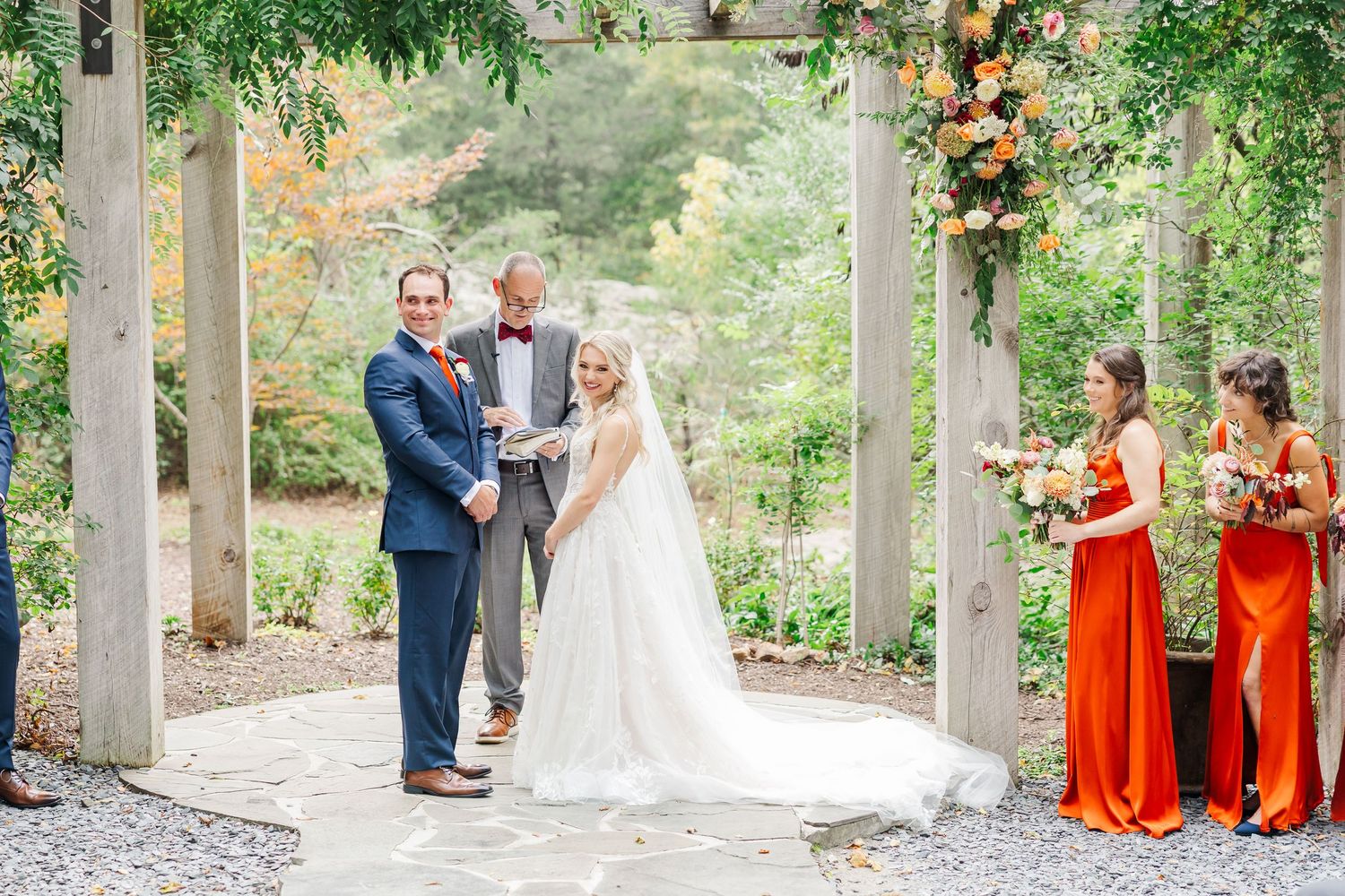 A wedding ceremony takes place under a floral-decorated pergola with bridesmaids in vibrant orange dresses.